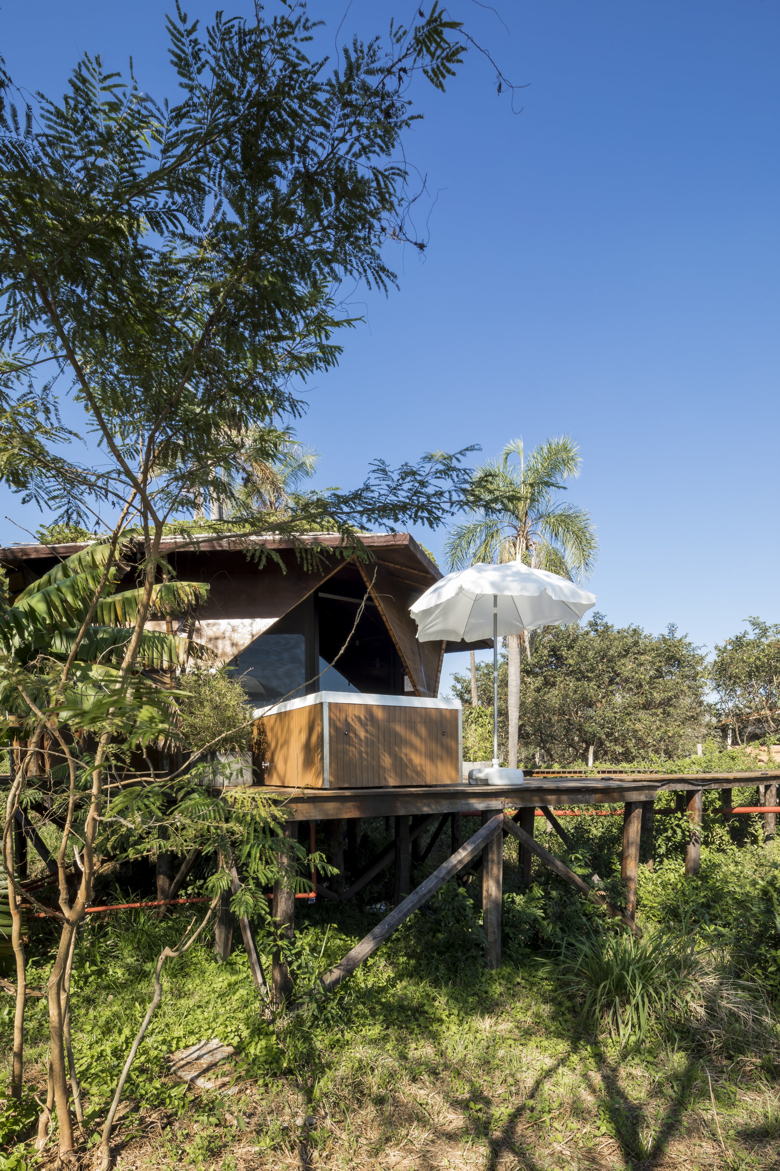 PRO ARQUITECTURA PARAGUAY architecture design PROSA
Casa de madera elevada sobre pilotes rodeada de vegetación y árboles, con una sombrilla blanca y cielo despejado.