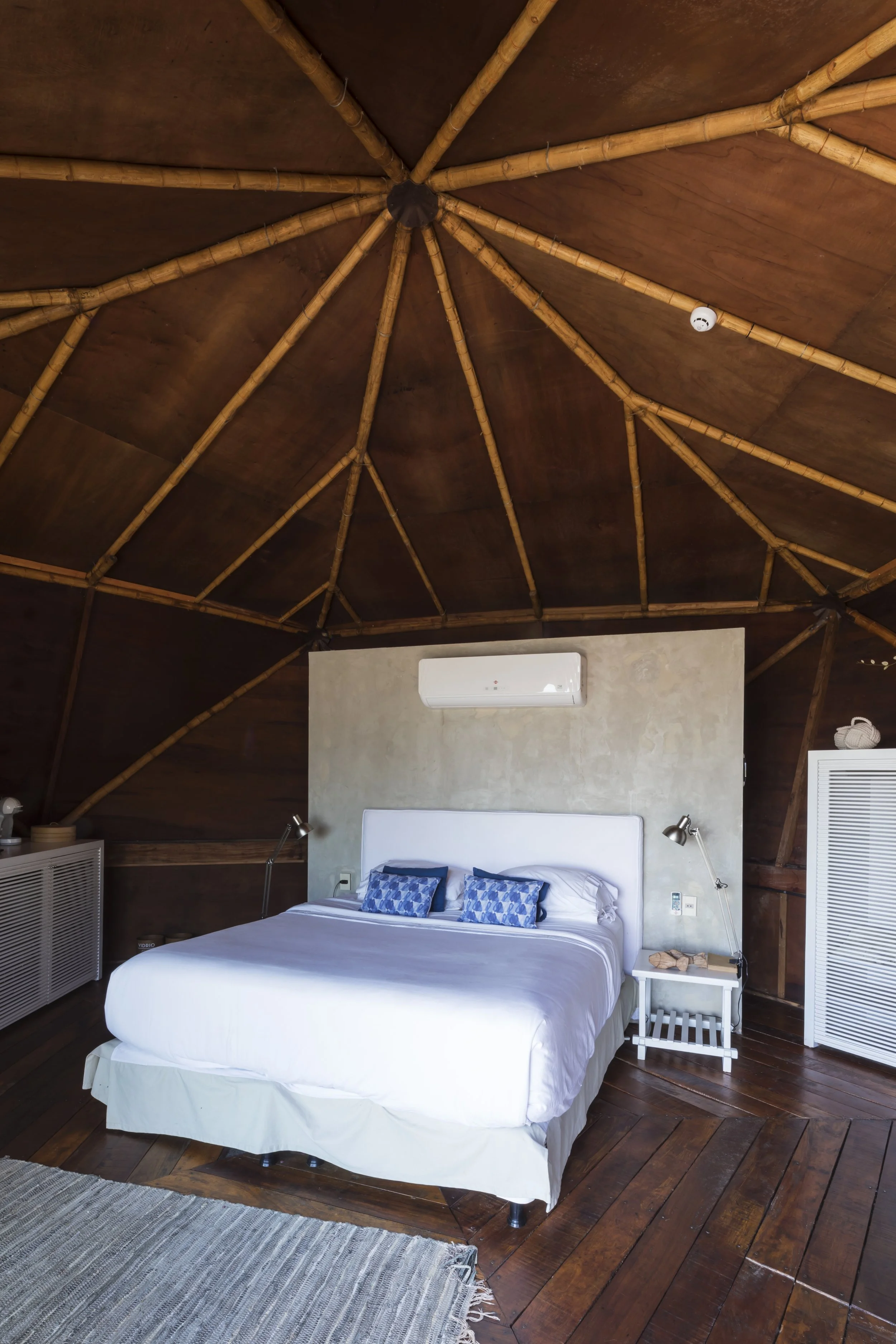 PRO ARQUITECTURA PARAGUAY architecture design PROSA
Habitación con techo de madera en forma de cúpula, cama matrimonial con cabecera blanca, almohadas azules, aire acondicionado y lámparas de lectura a los lados.
