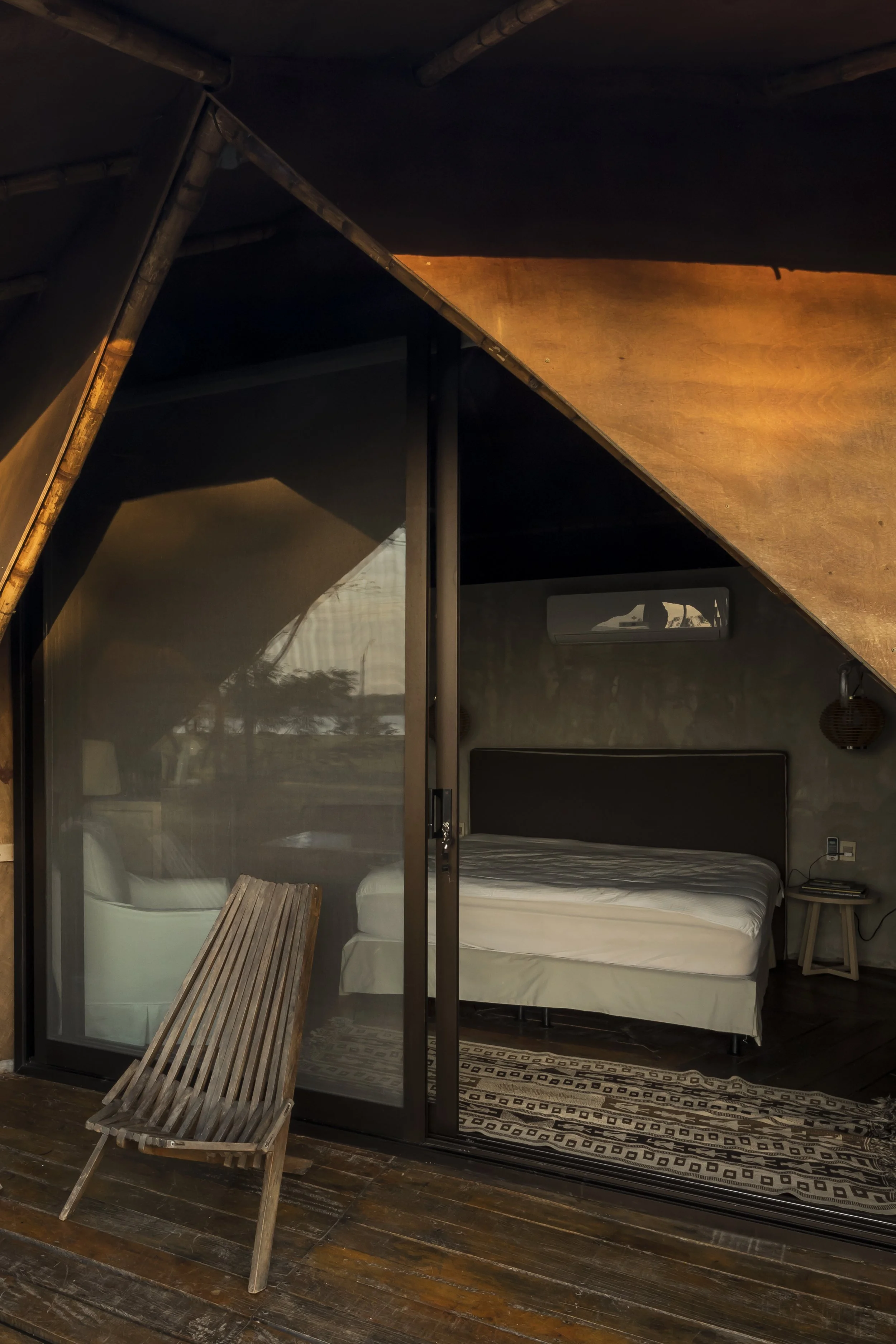 PRO ARQUITECTURA PARAGUAY architecture design PROSA
Vista de una habitación en una cabaña con paredes de madera y techo inclinado, con una cama y un sillón en la terraza exterior.