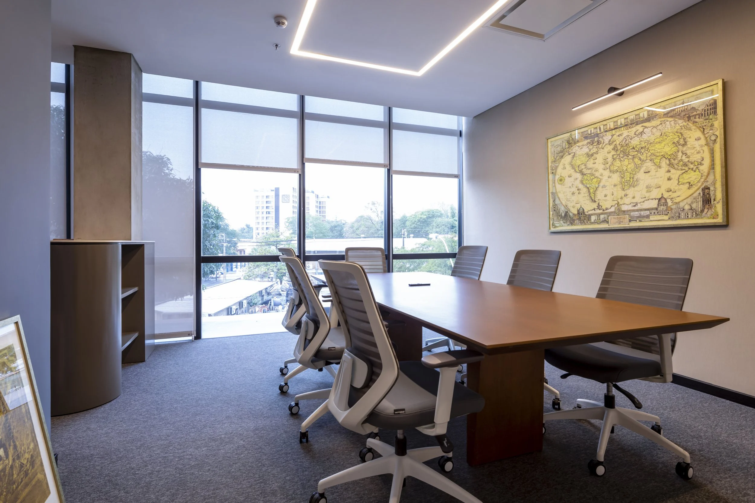 PRO ARQUITECTURA PARAGUAY architecture design PROSA
Sala de reuniones moderna con mesa de madera y sillas ergonómicas con ruedas, grandes ventanas que dejan entrar luz natural, cuadro de un mapa antiguo en la pared y una estantería pequeña.
