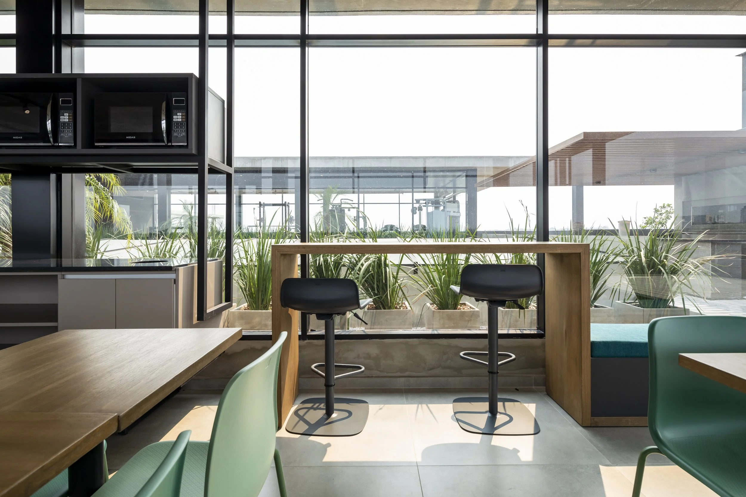 PRO ARQUITECTURA PARAGUAY architecture design PROSA
Interior de un café u oficina con mesas de madera y sillas verdes y negras, grandes ventanas con vista a plantas y un espacio exterior con equipo de ejercicio.