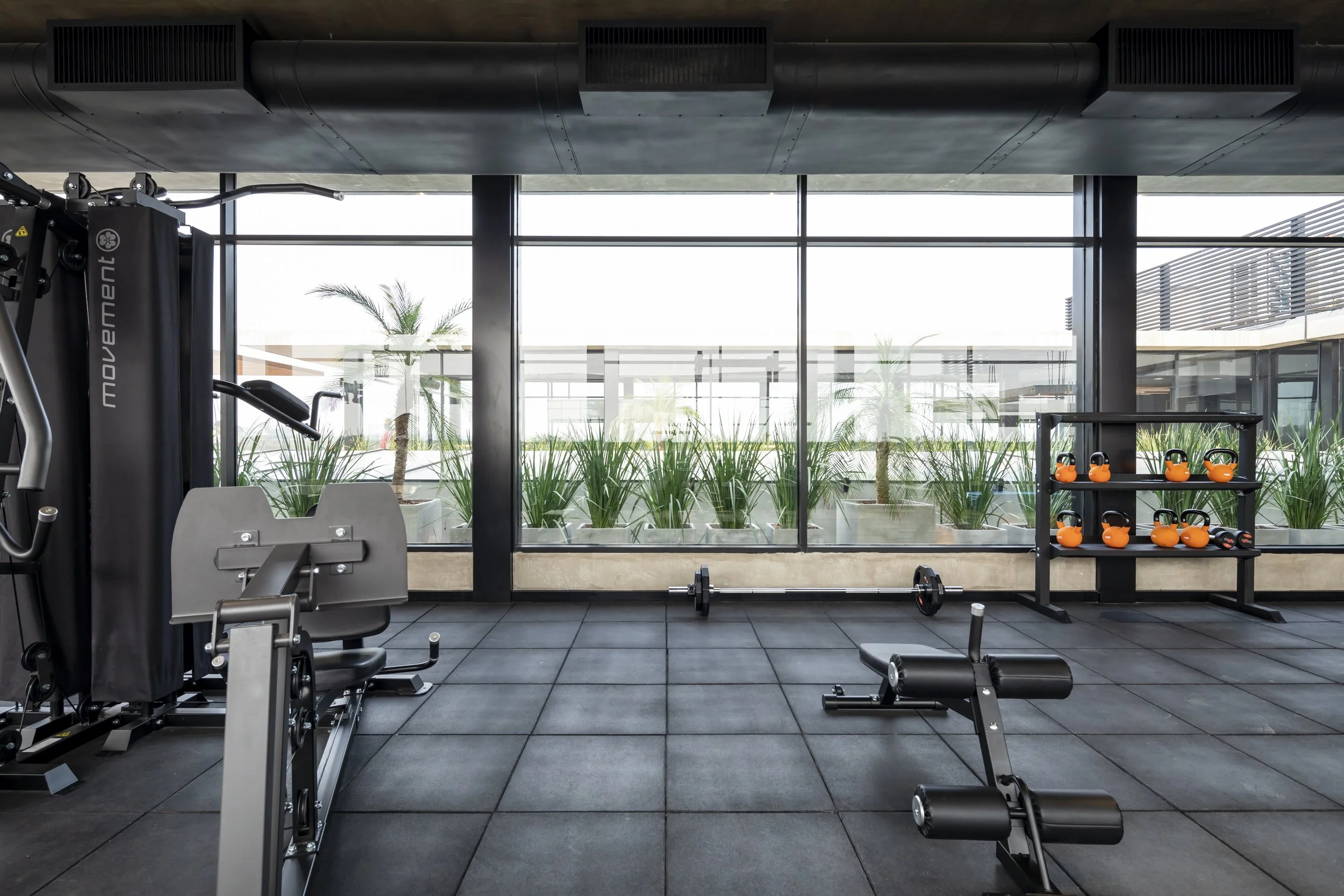 PRO ARQUITECTURA PARAGUAY architecture design PROSA
Área de gimnasio moderno con pisos de goma negra, máquinas de ejercicio, pesas y kettlebells de color naranja, amplio ventanal con vista a plantas y espacios exteriores