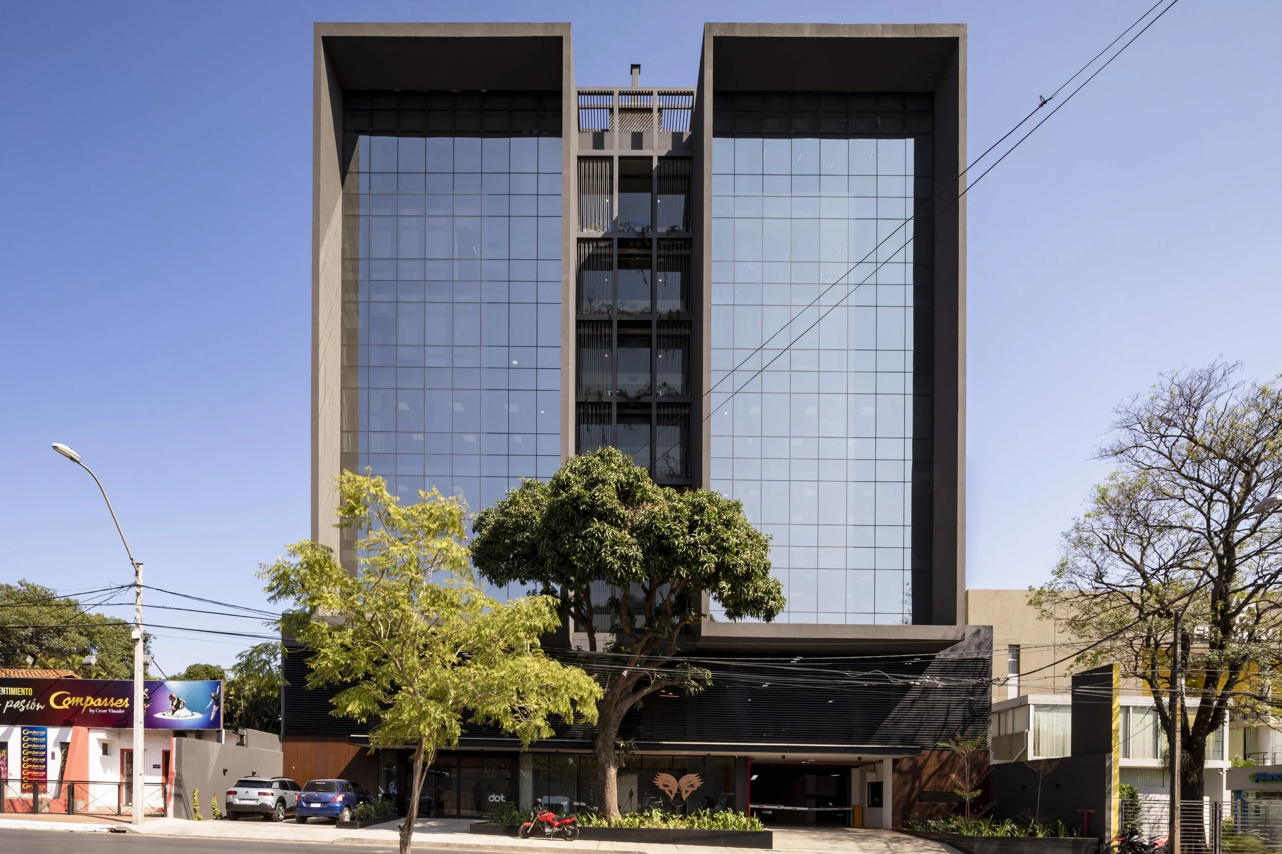 PRO ARQUITECTURA PARAGUAY architecture design PROSA
Edificio moderno de varios pisos con fachada de cristal y estructura de concreto, rodeado de árboles y con vehículos estacionados en la entrada.