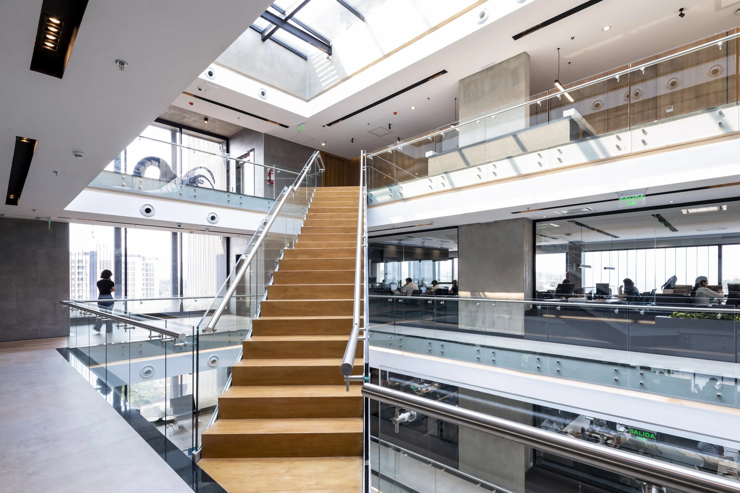 PRO ARQUITECTURA PARAGUAY architecture design PROSA
Interior de un edificio moderno con escaleras de madera y barandales de cristal, áreas de oficinas con empleados trabajando, paredes de cemento y ventanas grandes con vista a la ciudad.