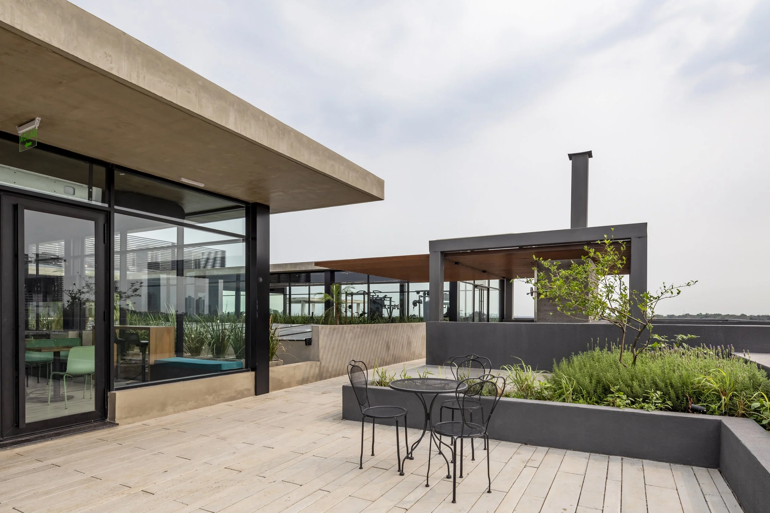 PRO ARQUITECTURA PARAGUAY architecture design PROSA
Terraza moderna con mesas y sillas negras, áreas verdes y vista urbana, edificio con grandes ventanales y estructura de concreto y madera.