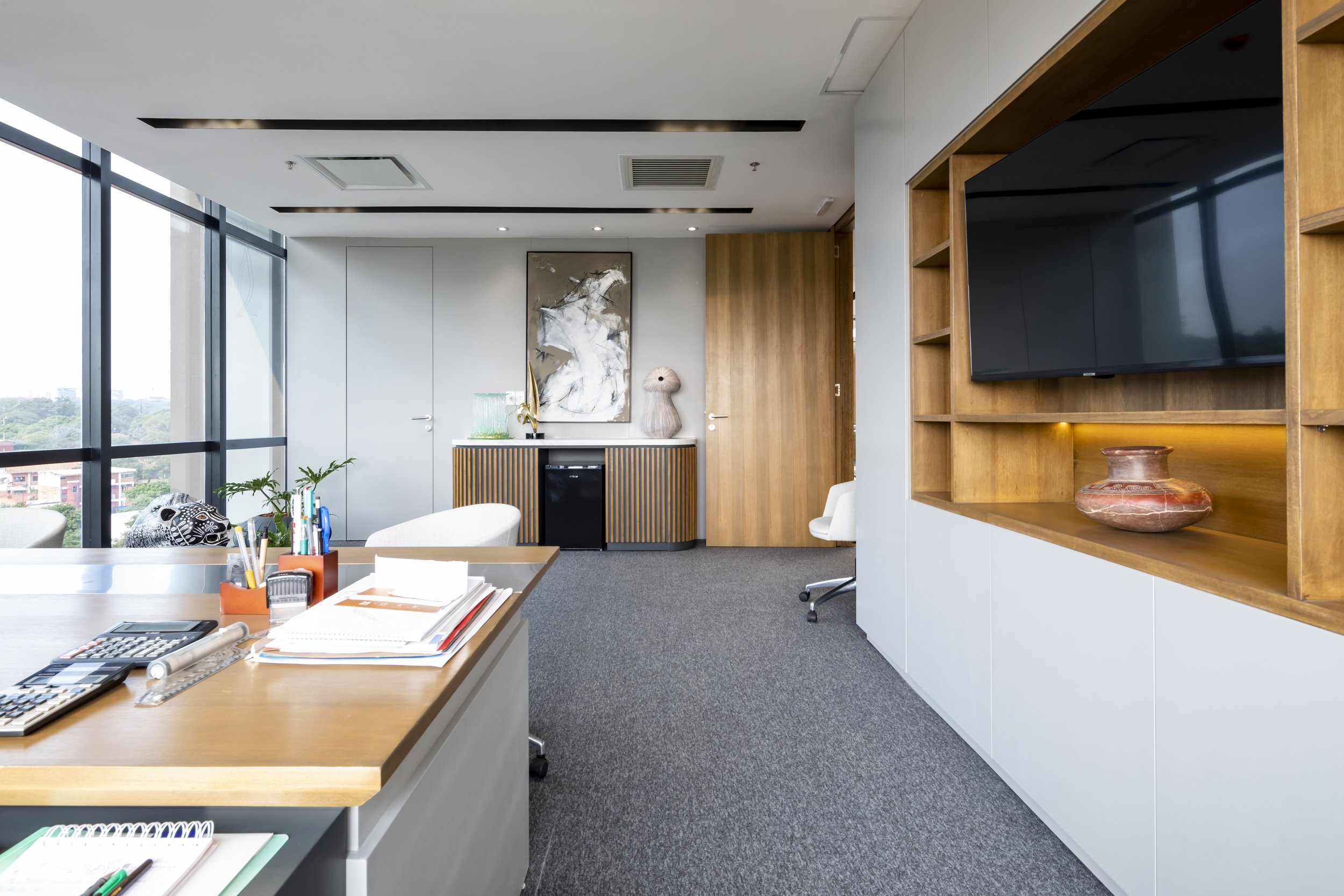 PRO ARQUITECTURA PARAGUAY architecture design PROSA
Oficina moderna con escritorio de madera, sillas blancas, estantería de madera con una televisión, decoraciones y vista a la ciudad por ventanas grandes.