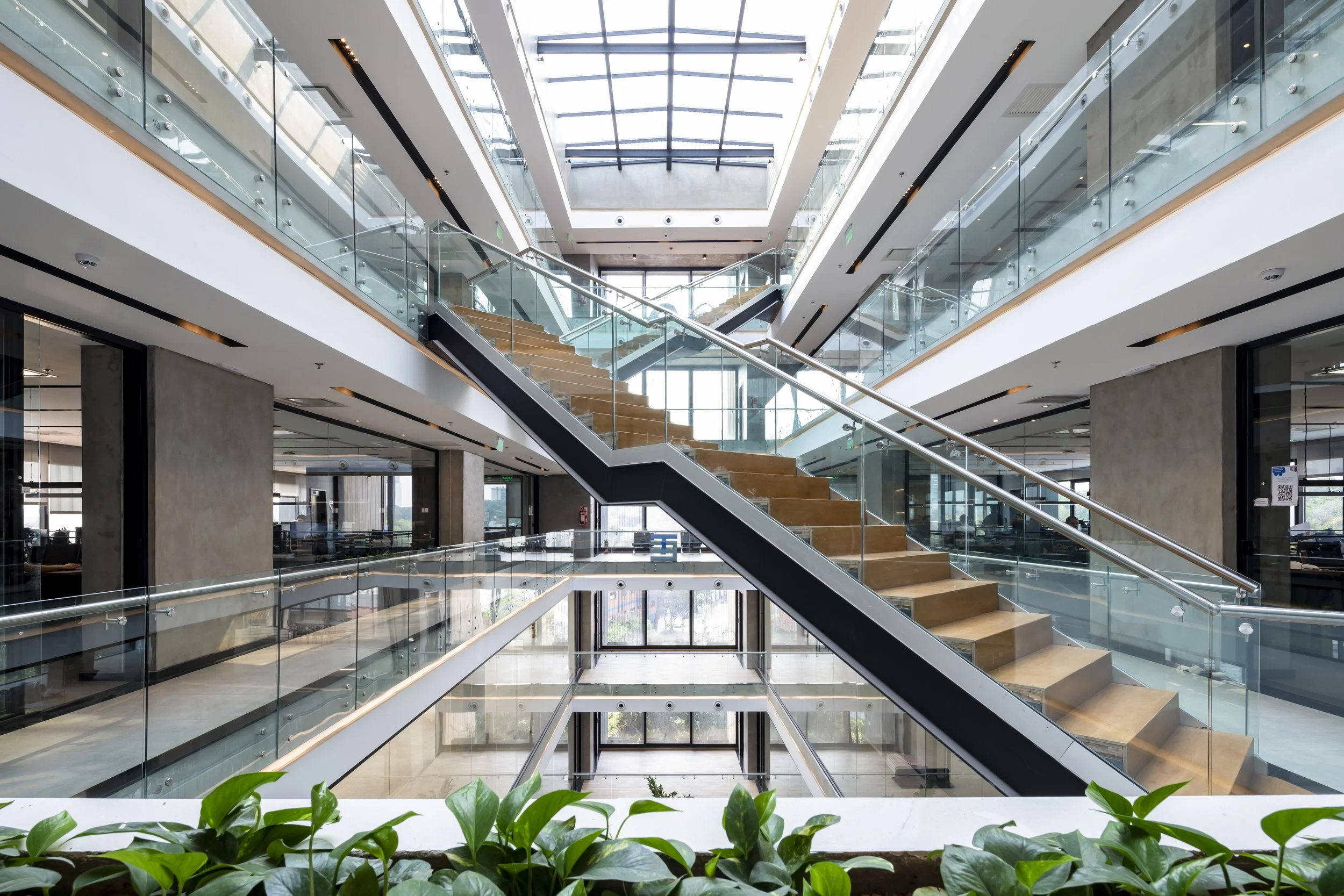PRO ARQUITECTURA PARAGUAY architecture design PROSA
Interior moderna de un edificio con escaleras de vidrio y madera, muchas ventanas de vidrio y plantas en primer plano.
