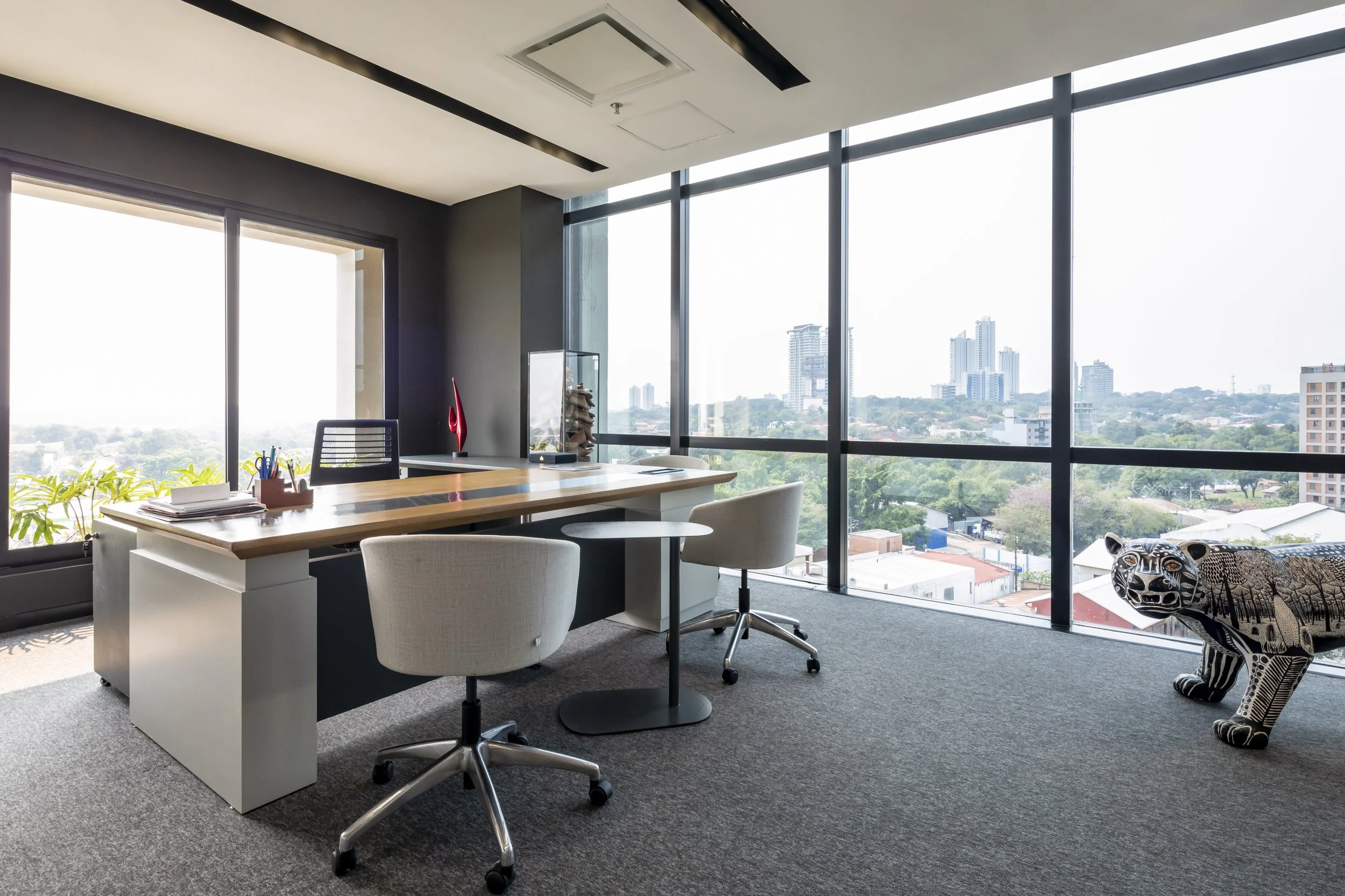 PRO ARQUITECTURA PARAGUAY architecture design PROSA
Una oficina moderna con vista a la ciudad, escritorio con sillas, lámpara, objetos decorativos y una escultura de un animal con diseño artístico.