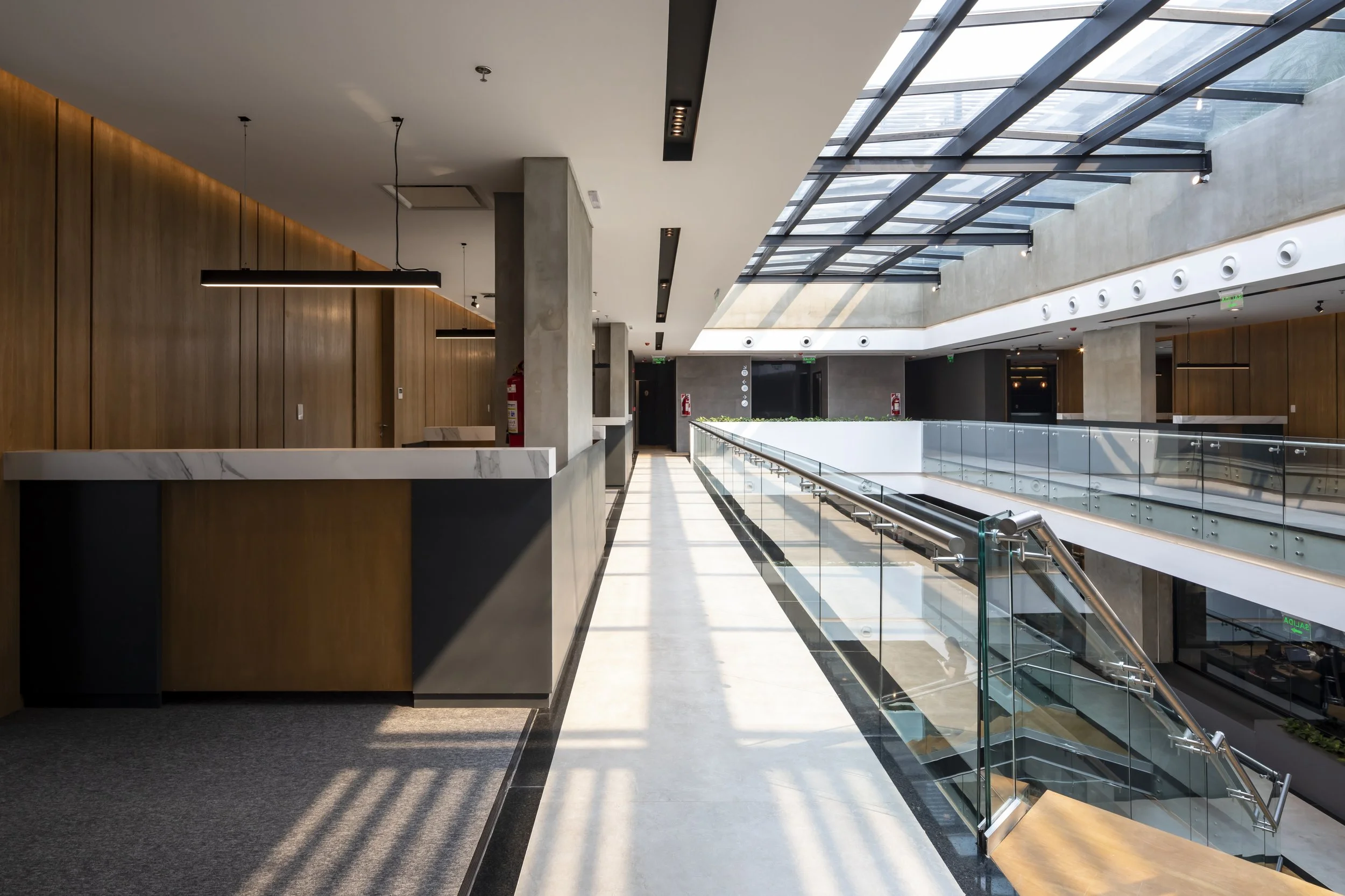 PRO ARQUITECTURA PARAGUAY architecture design PROSA
Pasillo de un edificio moderno con paredes de madera, un techo de vidrio, paredes de concreto y barandal de vidrio, con luz natural entrando por el techo.