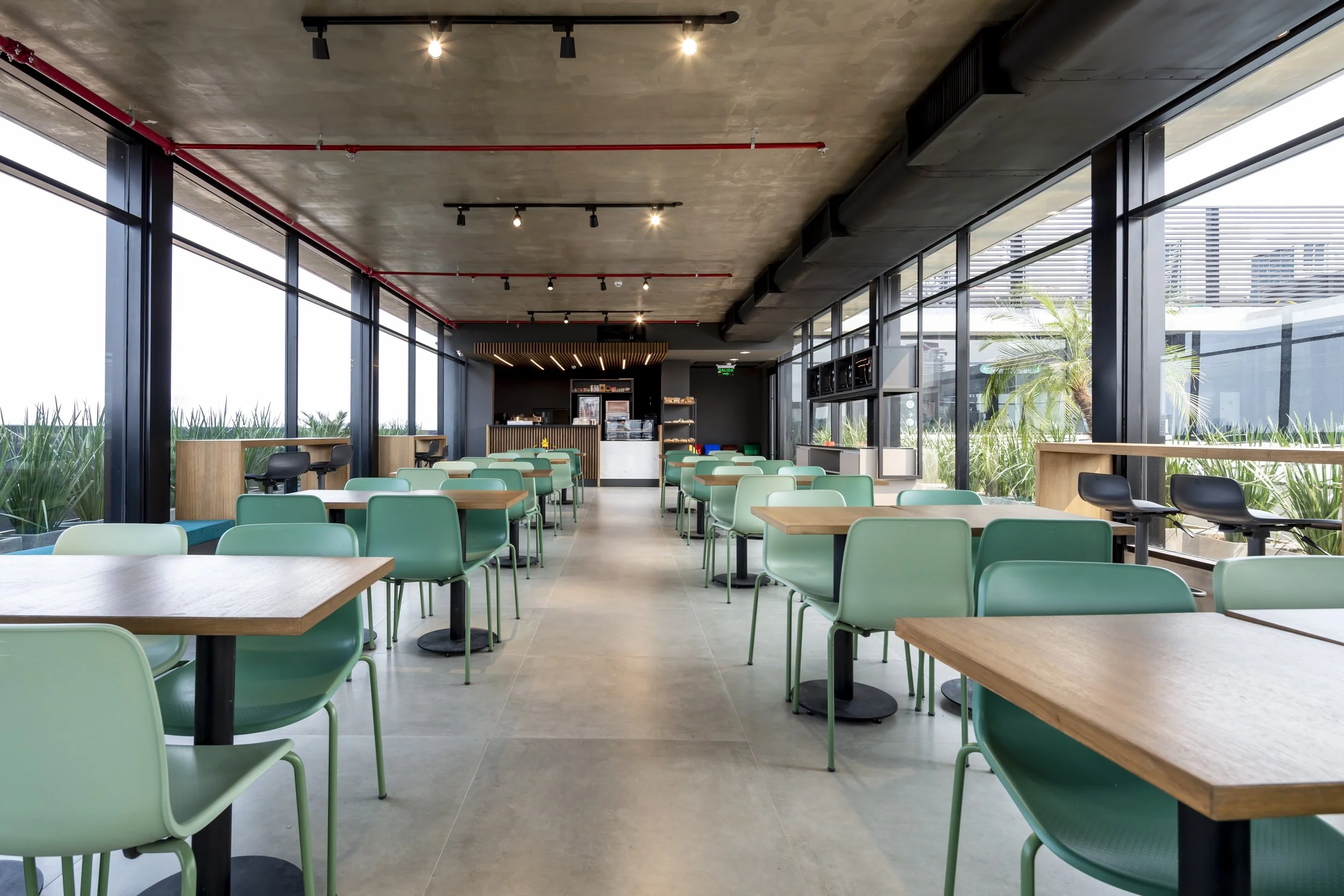 PRO ARQUITECTURA PARAGUAY architecture design PROSA
Interior de un restaurante moderno con mesas de madera y sillas verdes, grandes ventanales dejando ver plantas exteriores, iluminación artificial en el techo y un área de cafetería al fondo.