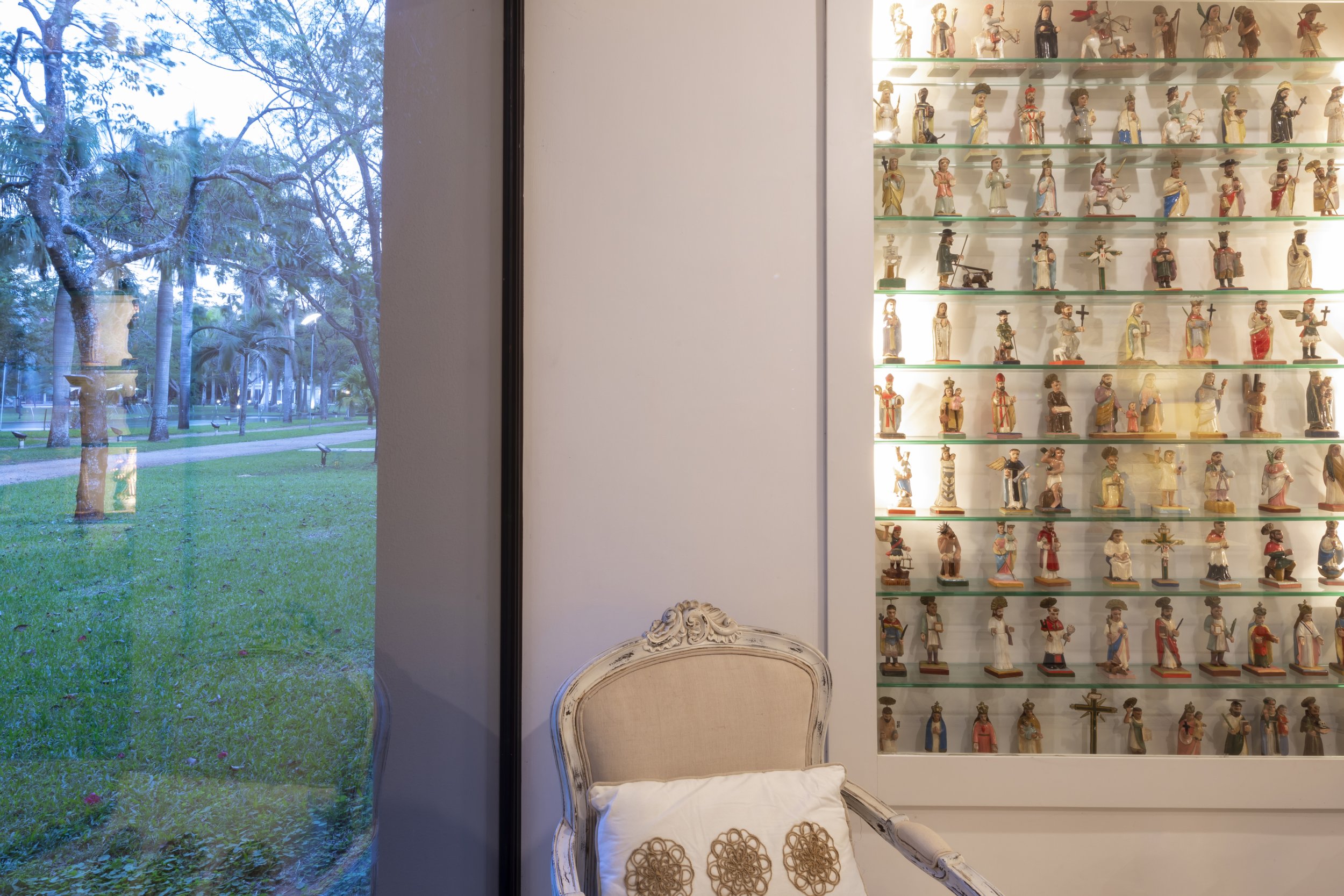 Interior de una habitación con silla antigua de madera blanca y cojín, y una pared con exhibición de figuras religiosas en vitrinas de vidrio, junto a una ventana que muestra un parque con árboles y césped.