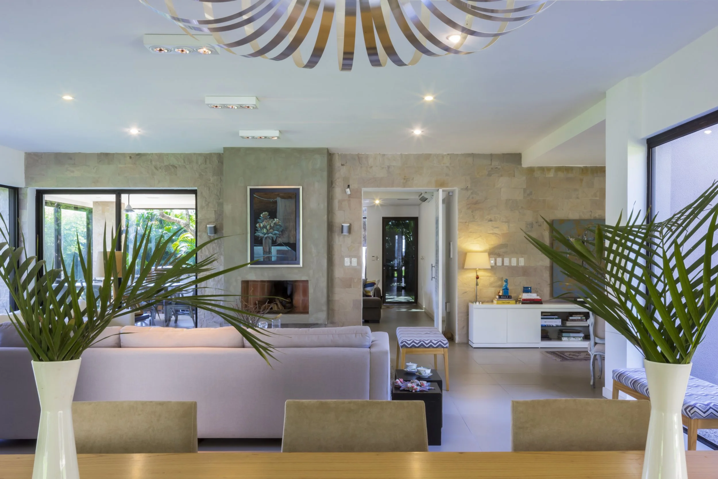 Sala moderna con sillones beige, plantas en jarrones blancos, chimenea, obras de arte en las paredes, lámparas de mesa, y ventanas grandes que dejan entrar luz natural.