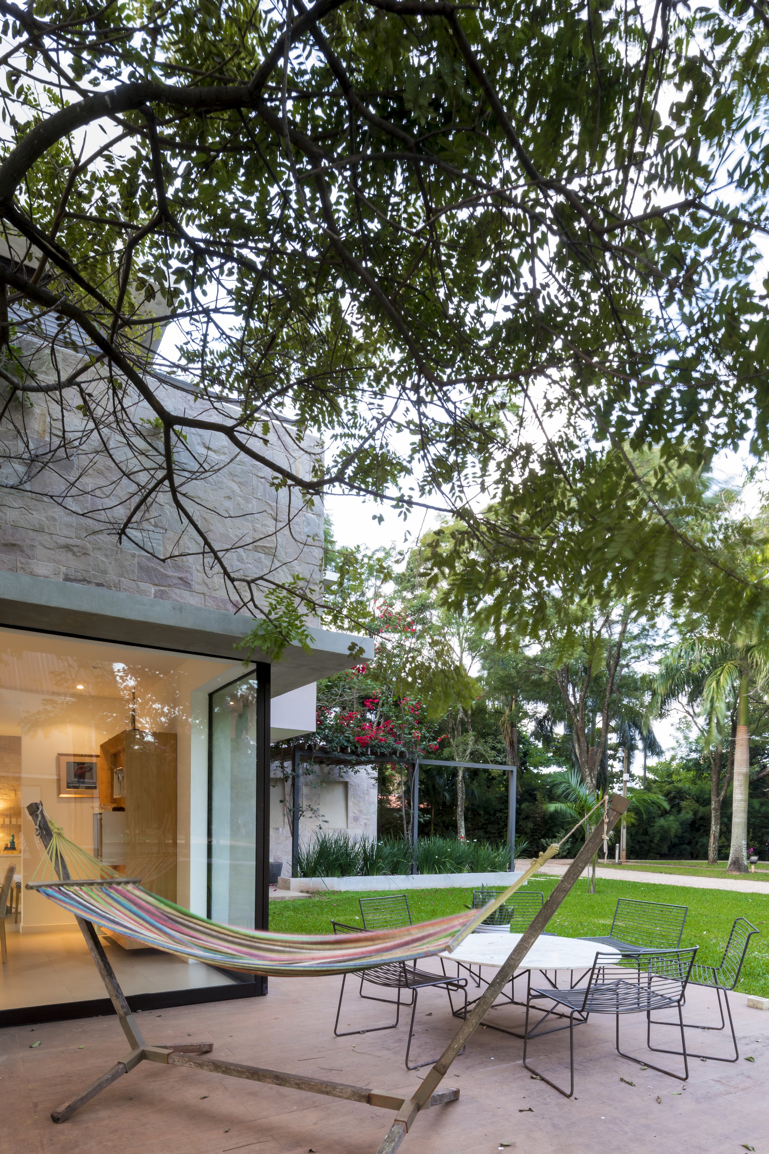 PRO ARQUITECTURA PARAGUAY architecture design PROSA
Espacio exterior con silla mecedora de tela en un soporte de madera, mesa de metal con varias sillas, césped y árboles en el fondo, y parte de la casa con ventanas de cristal.