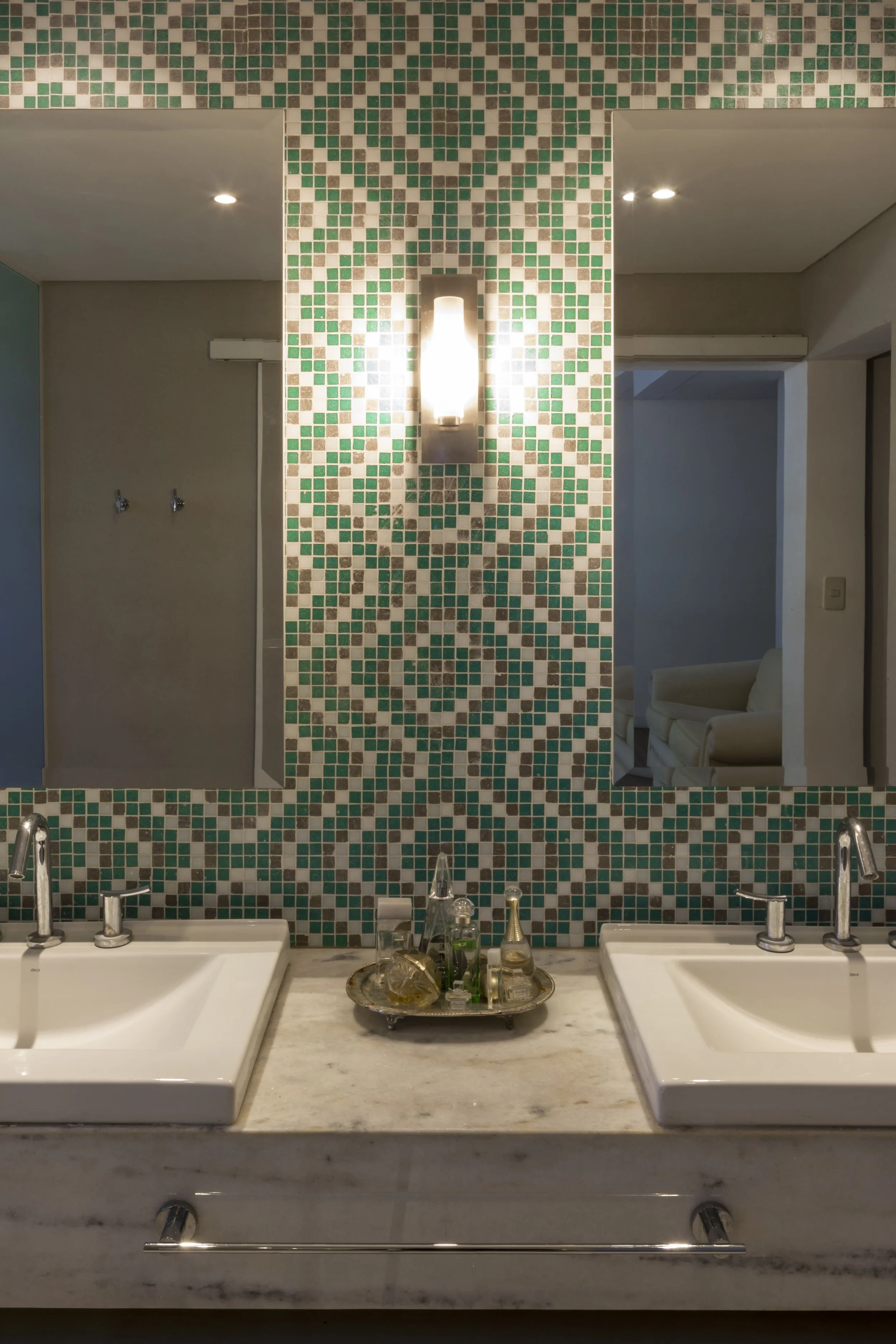 Espejo con mosaicos en tonos verdes y beige, con dos lavabos de mármol y decoración en la encimera, en un baño. Se refleja una pared con lámpara en el espejo.