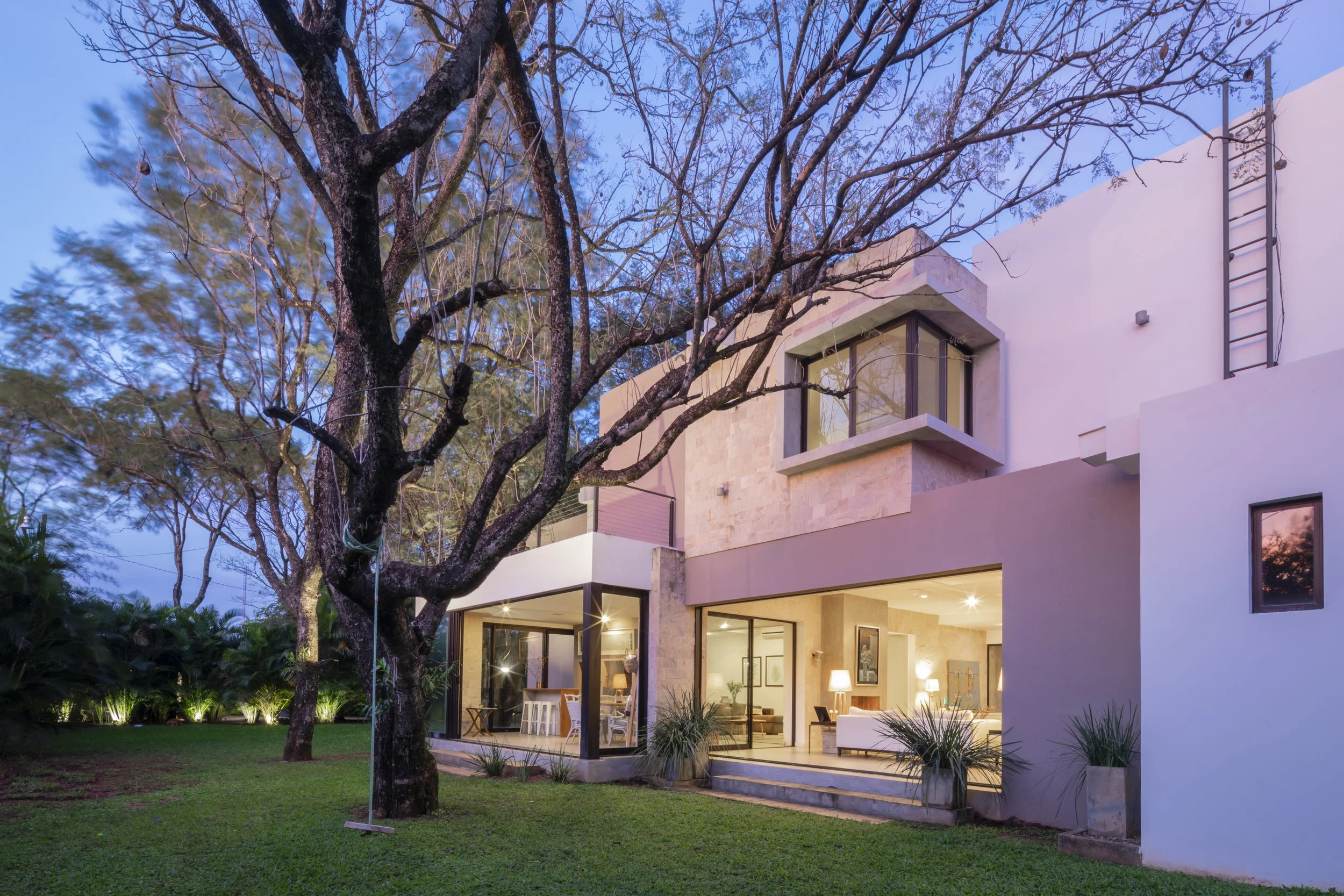PRO ARQUITECTURA PARAGUAY architecture design PROSA
Casa moderna de dos niveles con paredes blancas y ventanas grandes, iluminada en la noche, con un árbol grande en el jardín y un columpio de cuerda colgado de sus ramas.