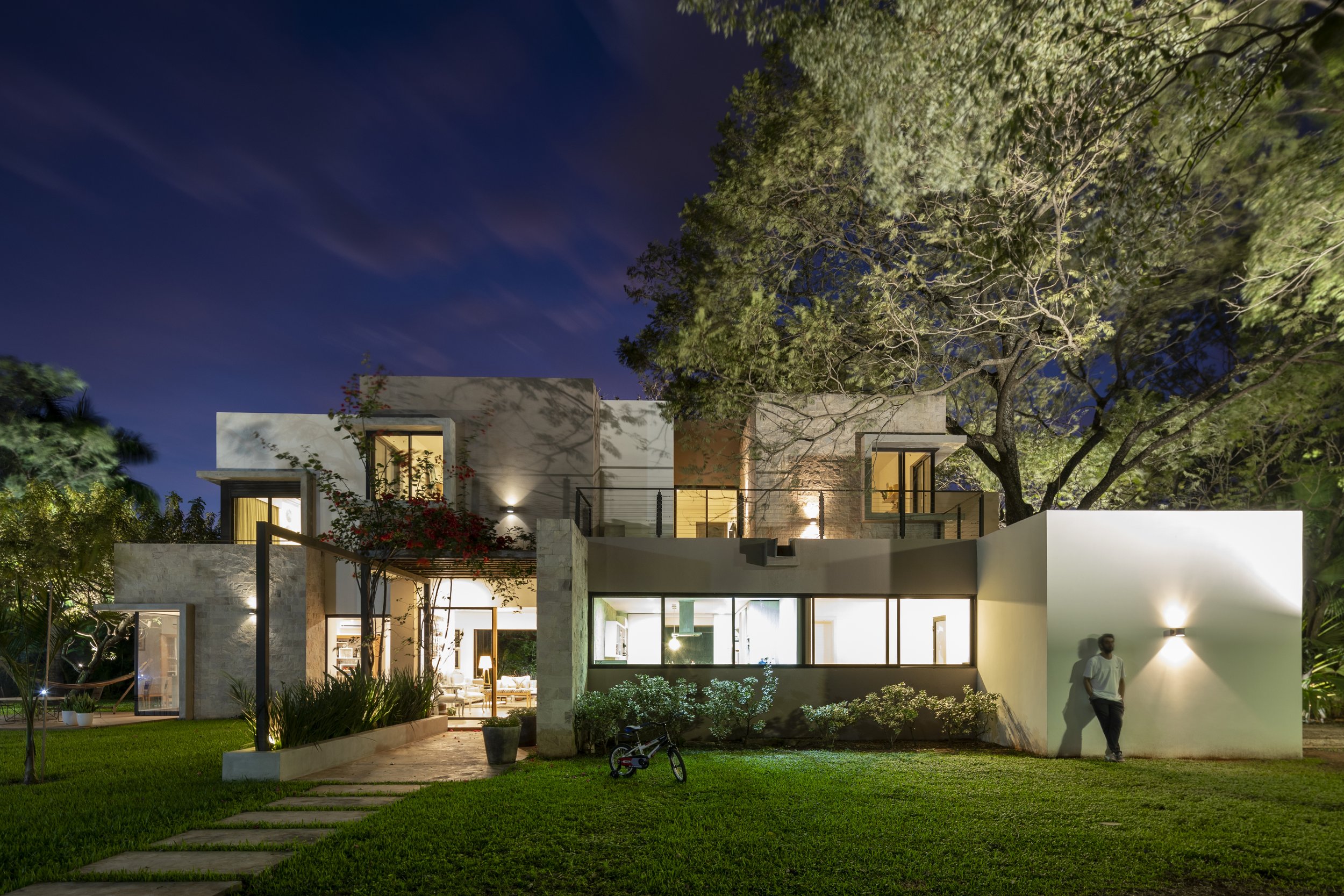 PRO ARQUITECTURA PARAGUAY architecture design PROSA
Casa moderna iluminada por la noche con árboles y césped en el jardín, una persona apoyada en la pared y una bicicleta en el césped.