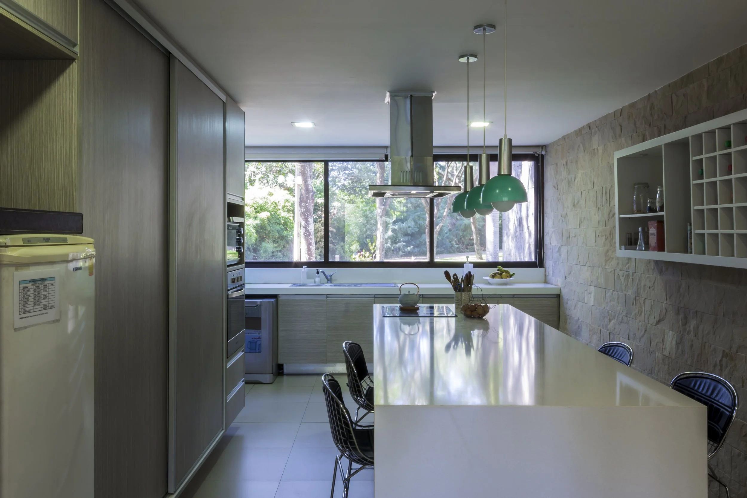 Cocina moderna con mesón de granito y sillas de metal, campana extractora, ventanas grandes con vista a árboles, paredes de piedra y estanterías blancas con utensilios.