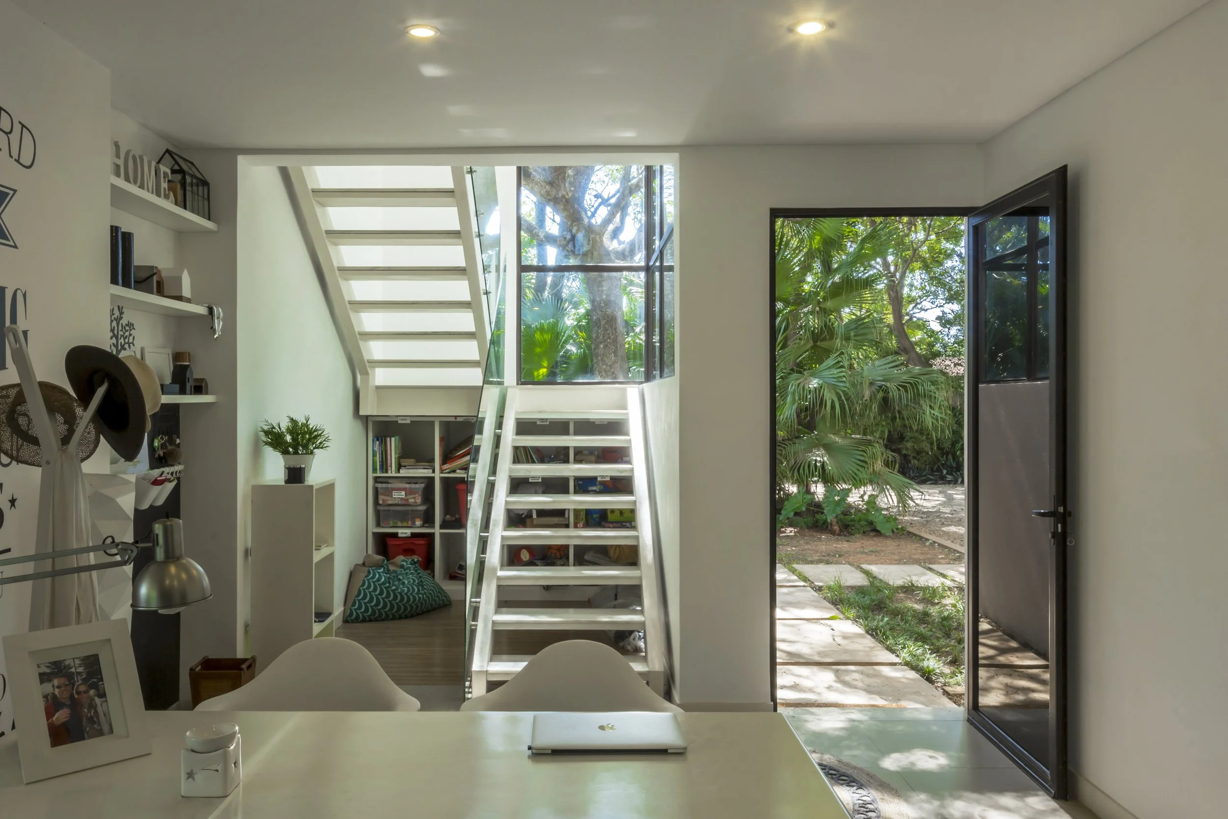 PRO ARQUITECTURA PARAGUAY archiInterior de una casa con escalera blanca, puerta abierta que muestra un jardín con plantas verdes y un camino de piedra, escritorio con una maceta y una laptop cerrada, estantes blancos con libros y objetos decorativos.