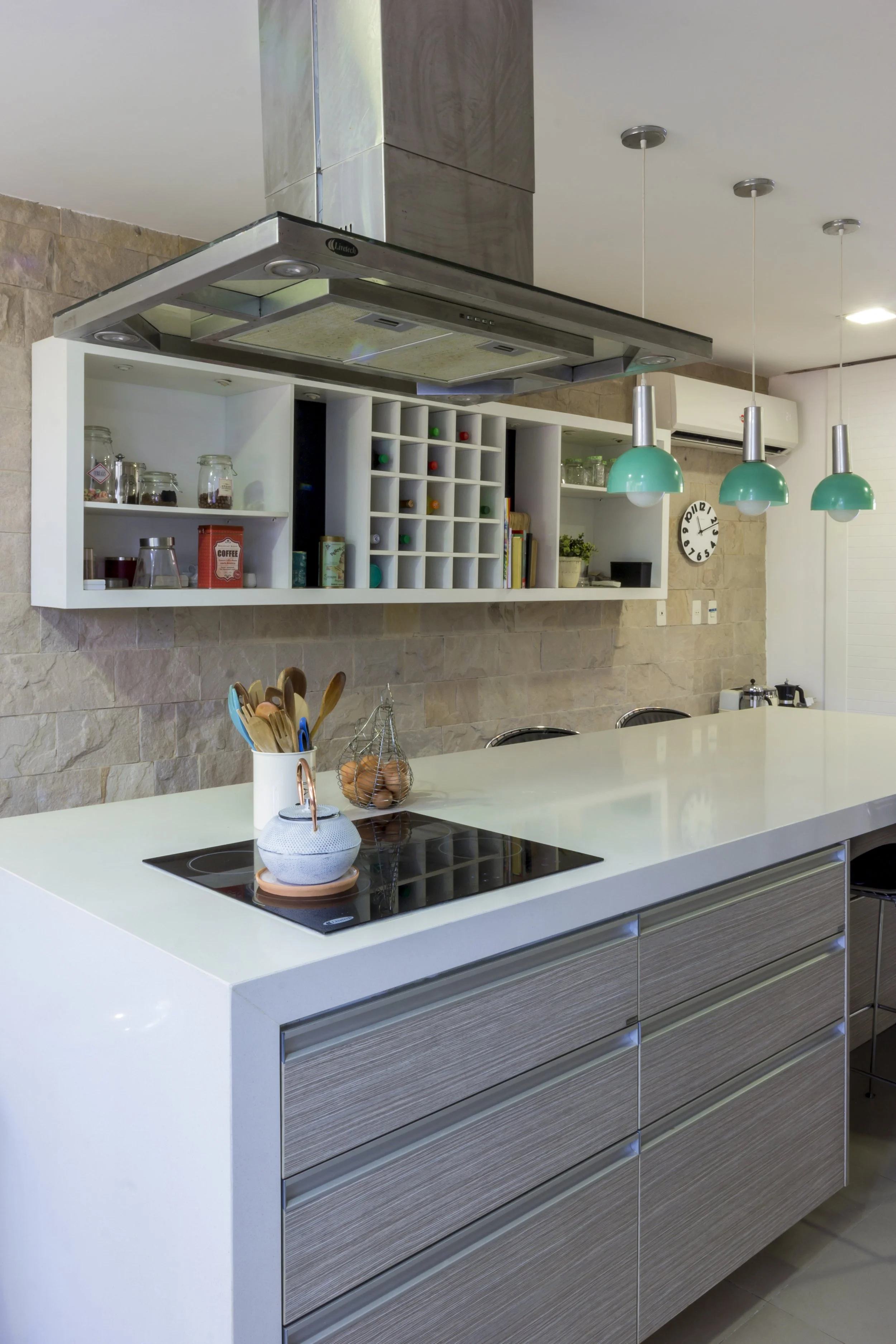 Vista de una cocina moderna con isla blanca, cajones de madera, una estufa eléctrica y una campana extractora de acero suspendida del techo. En el fondo, una pared de piedra y estanterías blancas con utensilios, libros y decoración.
