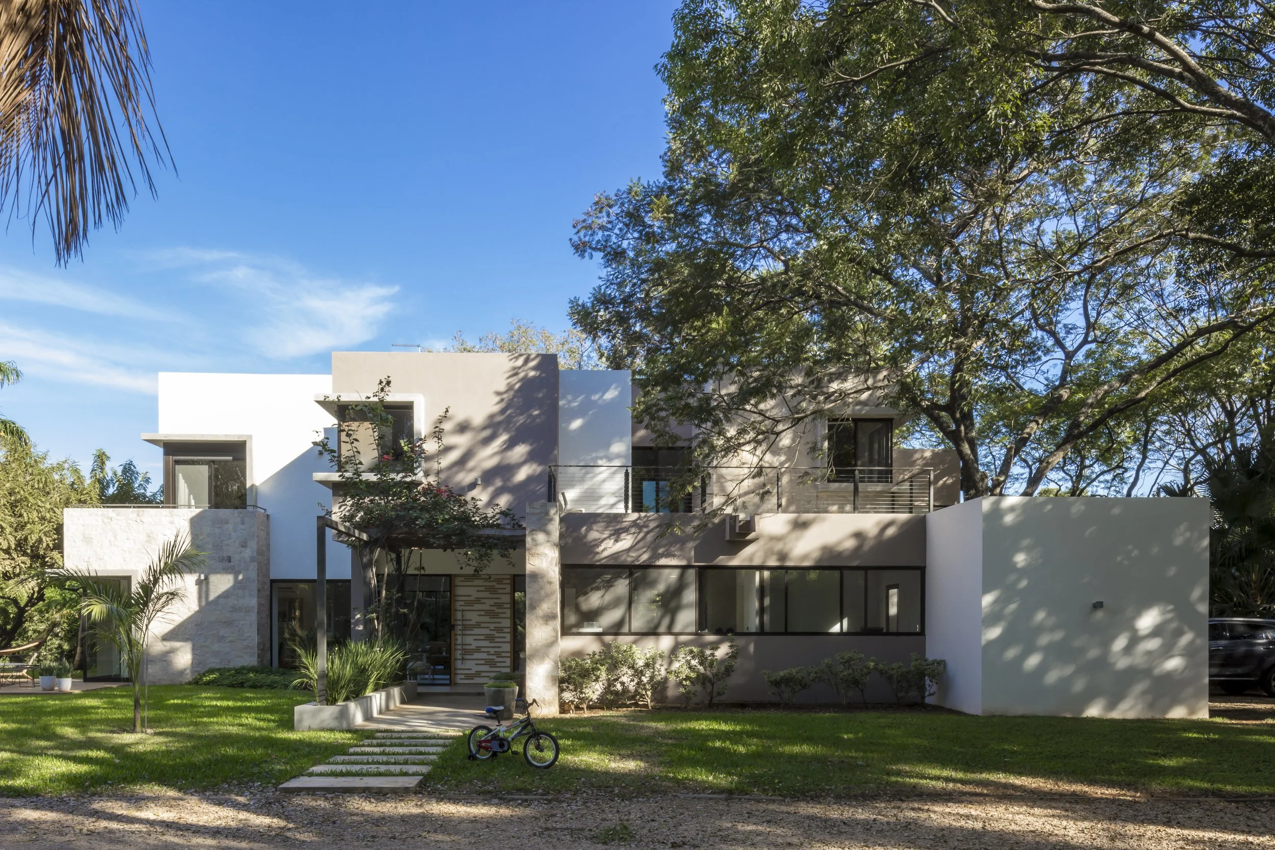 Casa moderna de dos pisos con paredes blancas y detalles de piedra, rodeada de árboles y césped, con bicicleta y camino de piedra en el jardín.