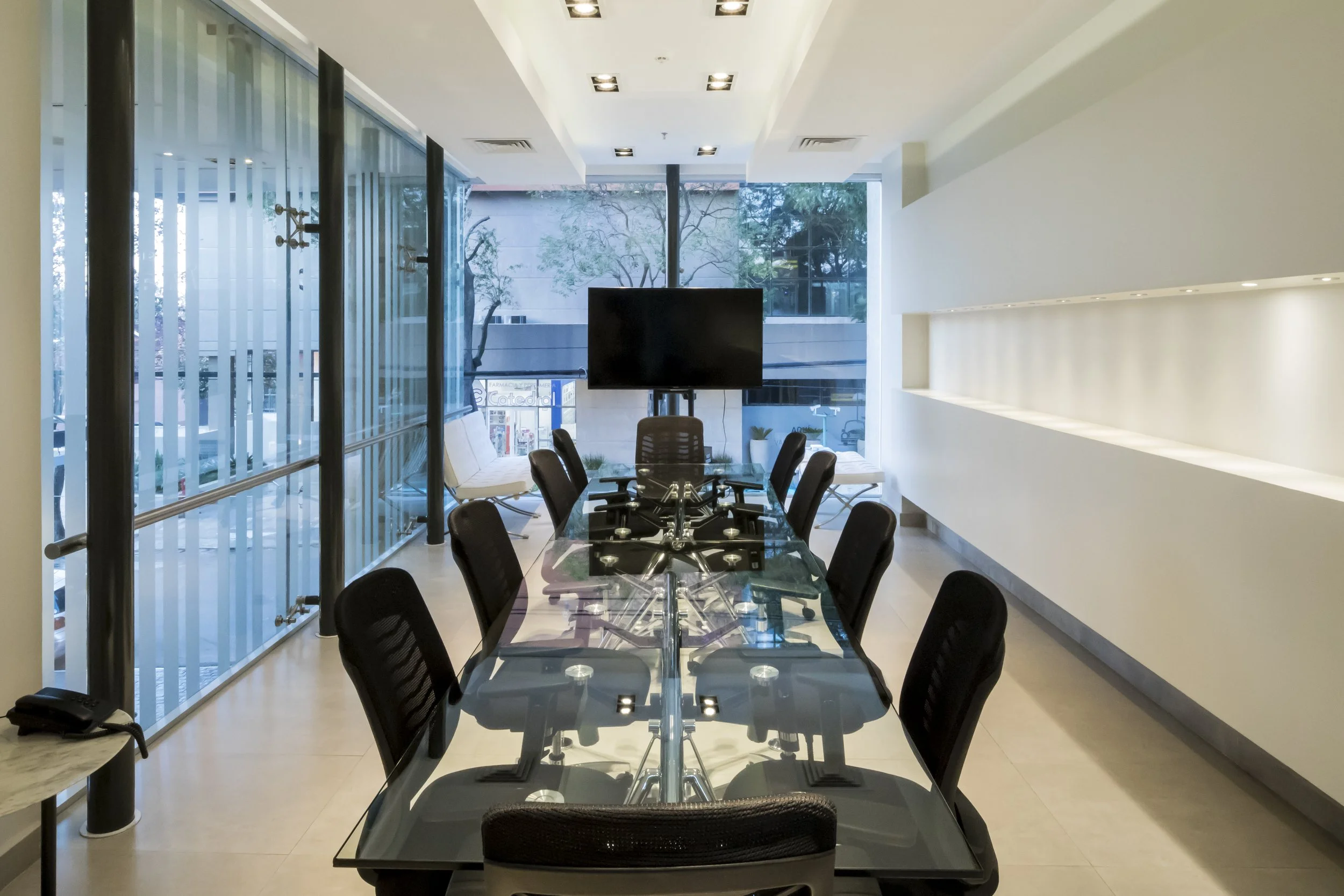 Sala de reuniones con mesa de vidrio y ocho sillas negras, pantalla plana en el fondo, ventanas grandes con vista a la calle, paredes blancas y iluminación moderna.