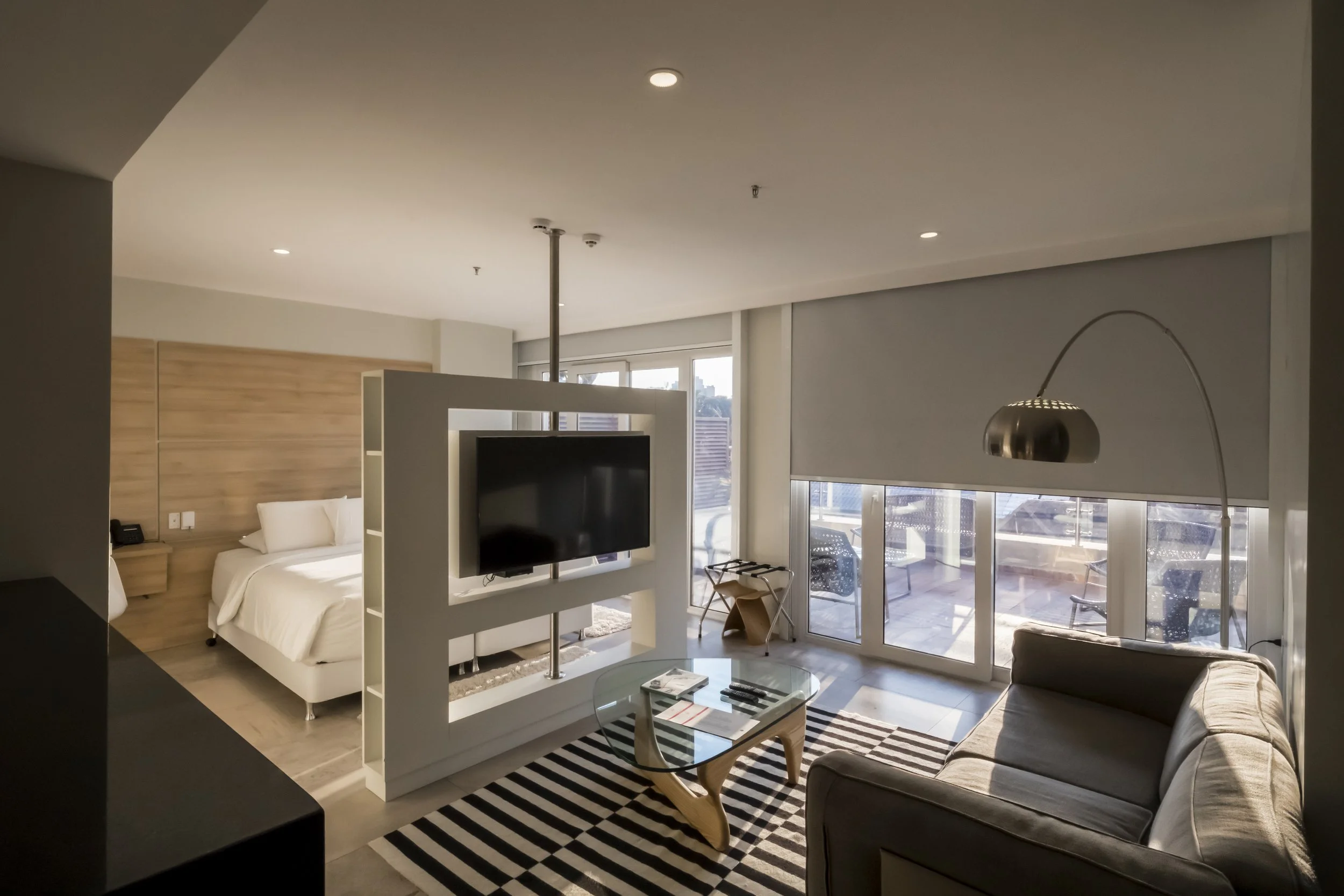 Sala de estar moderna con sillón gris, mesa de vidrio, alfombra a rayas, televisión, lámpara de arco, y balcón con sillones.