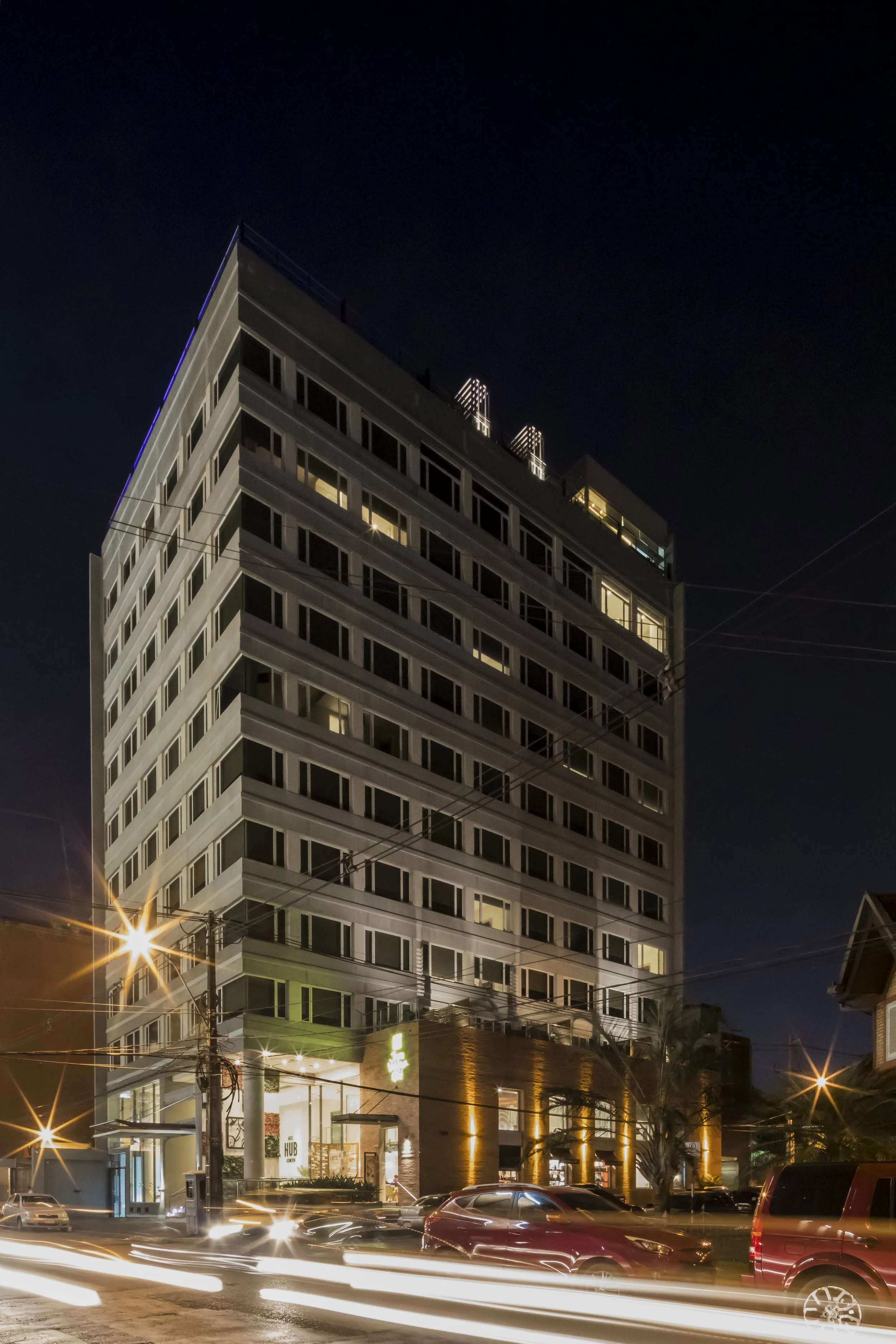 Edificio de varios pisos iluminado de noche, con luces en las ventanas y en la entrada principal, con autos circulando en la calle frente a él.