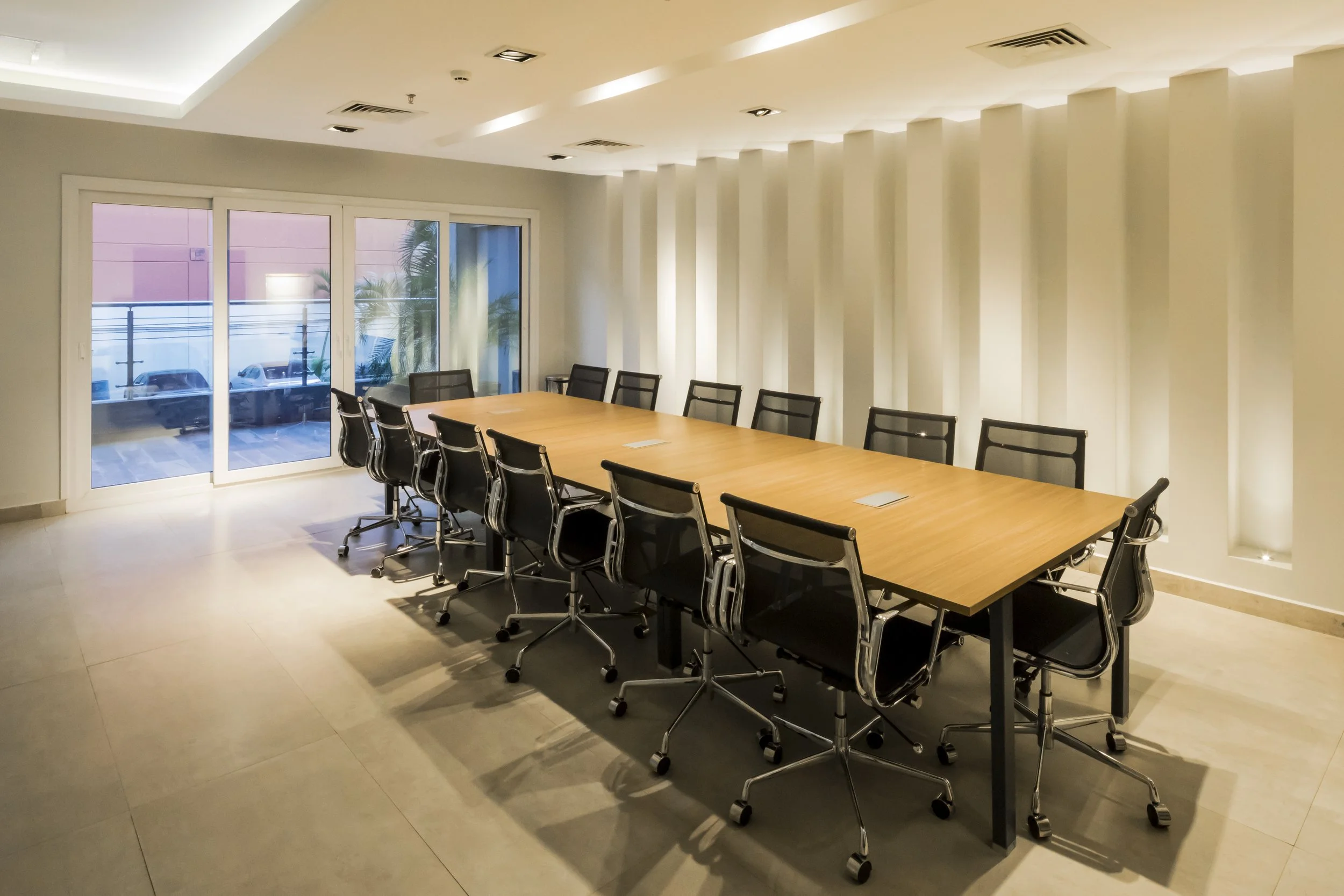 Sala de reuniones con mesa larga de madera y sillas negras con ruedas, ventanas grandes con vista a un balcón con autos y plantas, paredes blancas con iluminación moderna.