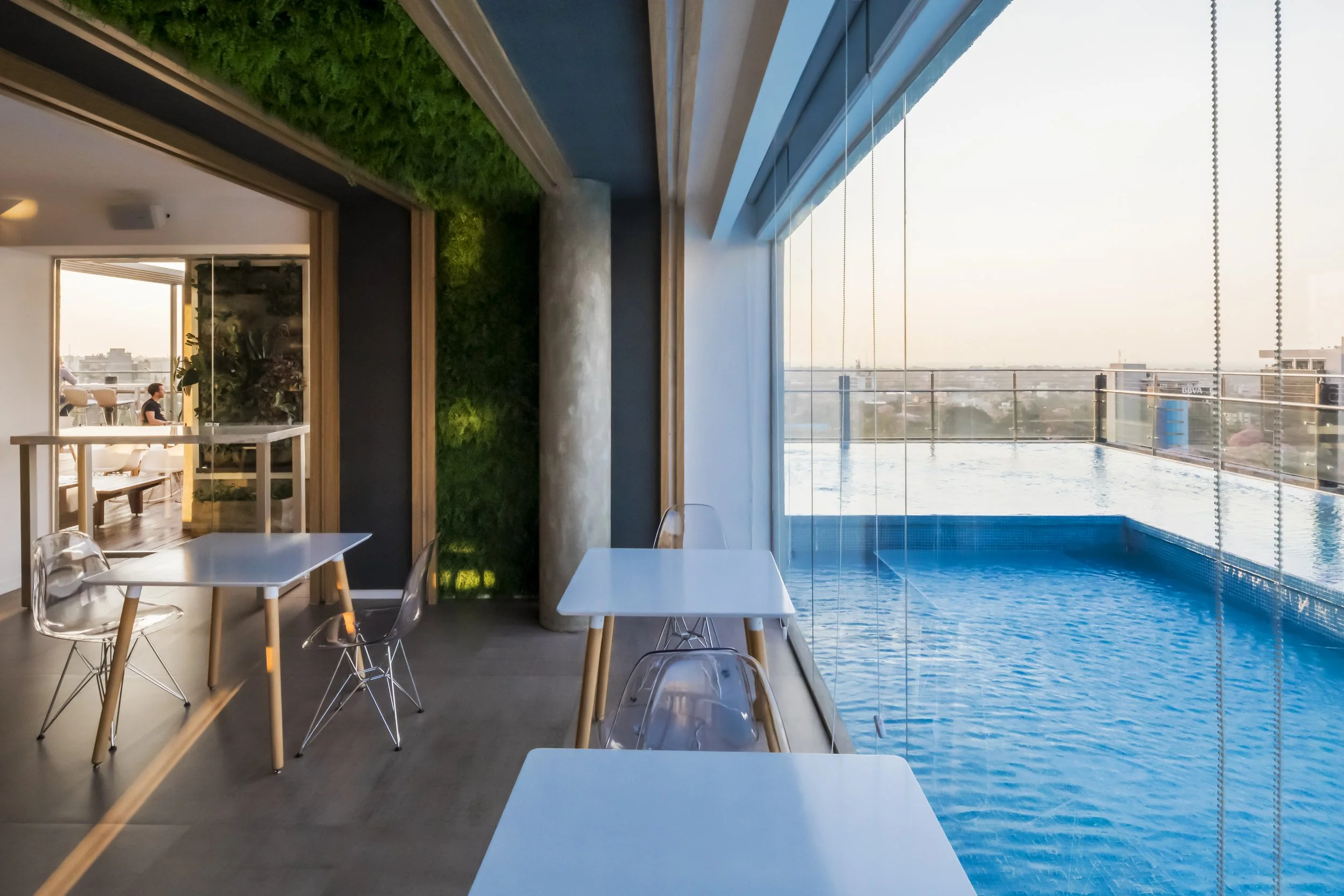Interior de un restaurante con vista a una alberca y la ciudad al atardecer, con mesas y sillas de diseño moderno, paredes decoradas con vegetación y grandes ventanas de vidrio.