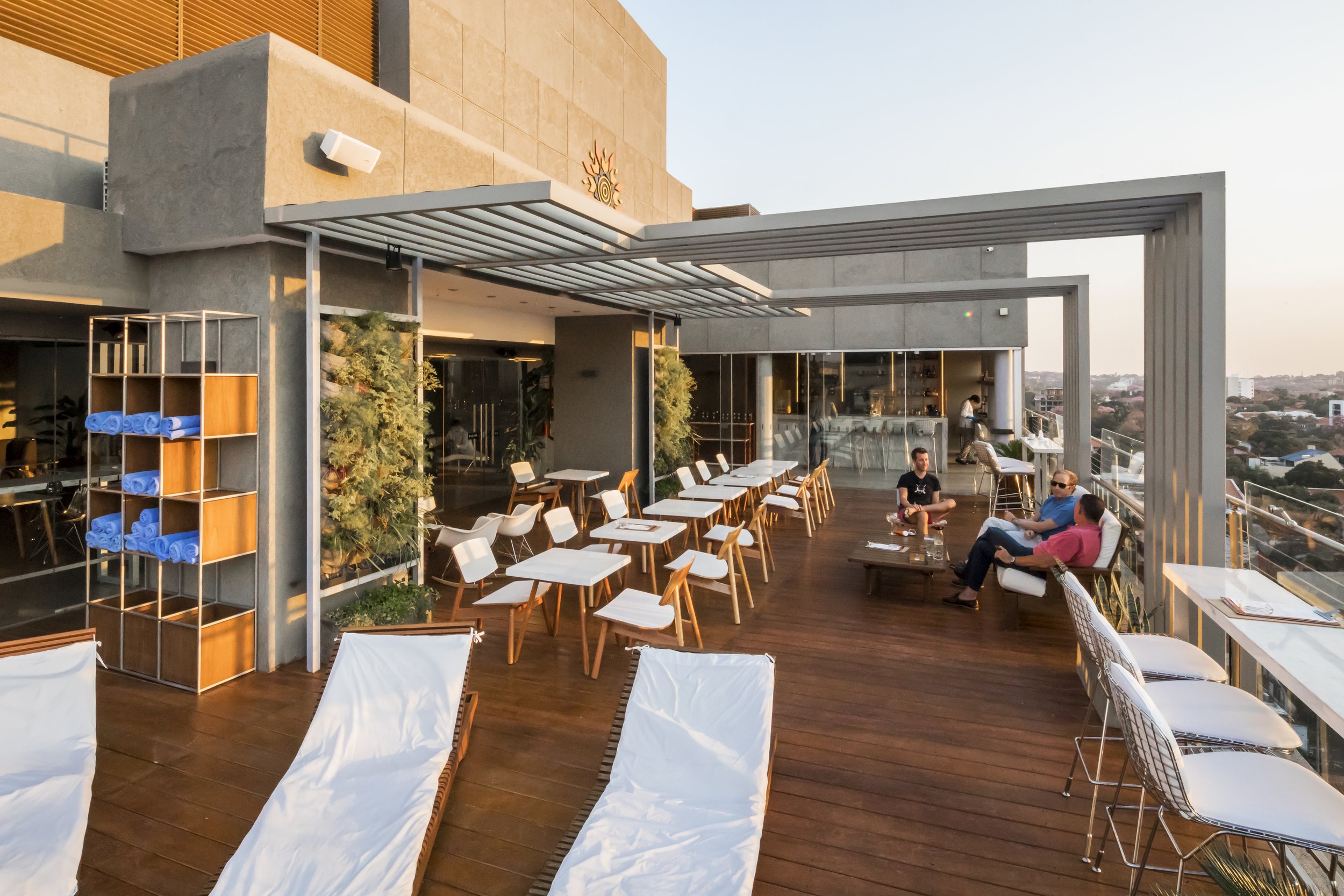 Terraza decorada moderna con sillones, mesas, sillas, tumbonas y plantas, con vista urbana de atardecer.