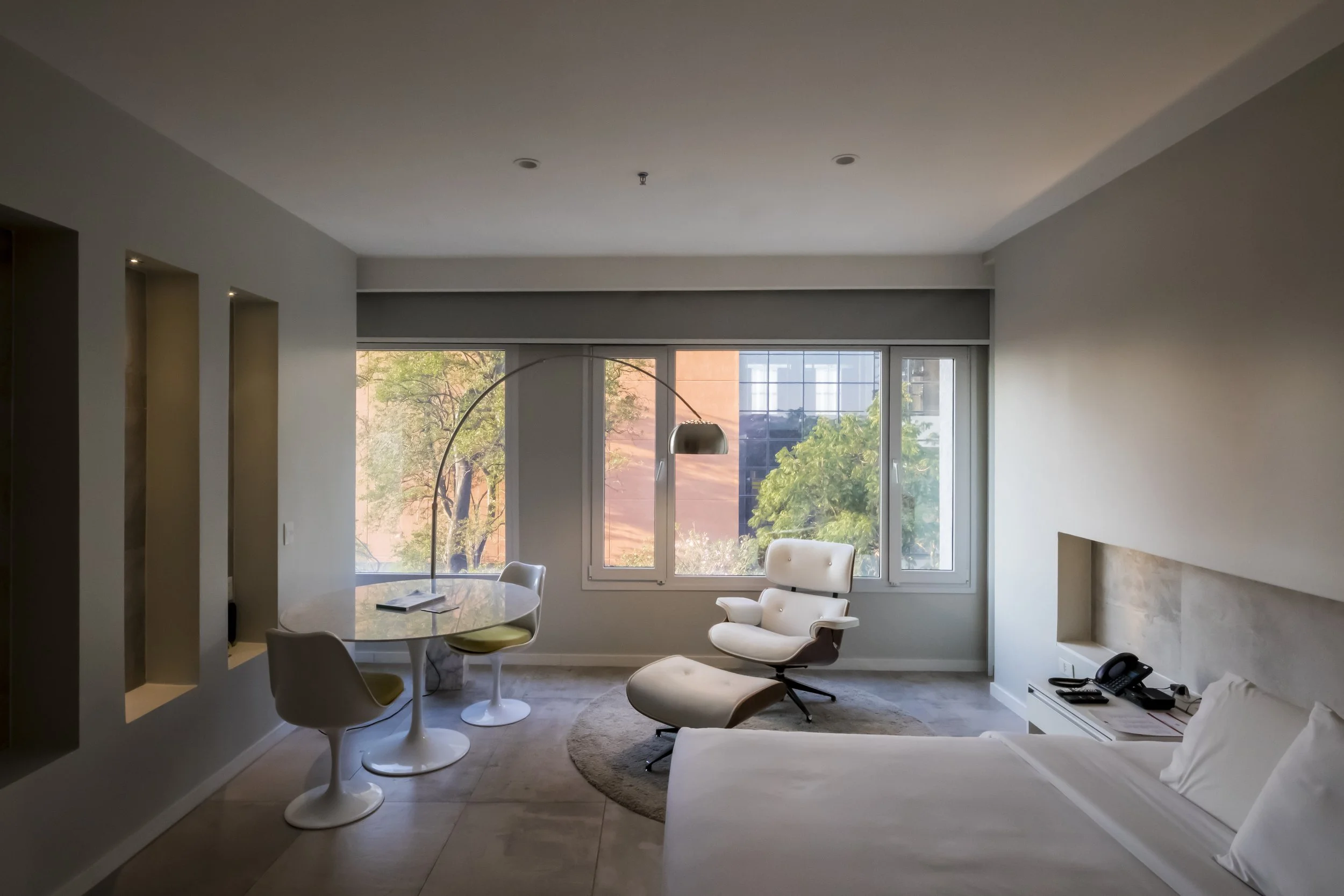 Sala moderna con sillón blanco, mesa redonda con sillas, lámpara arqueada y ventanas grandes con vista a árboles