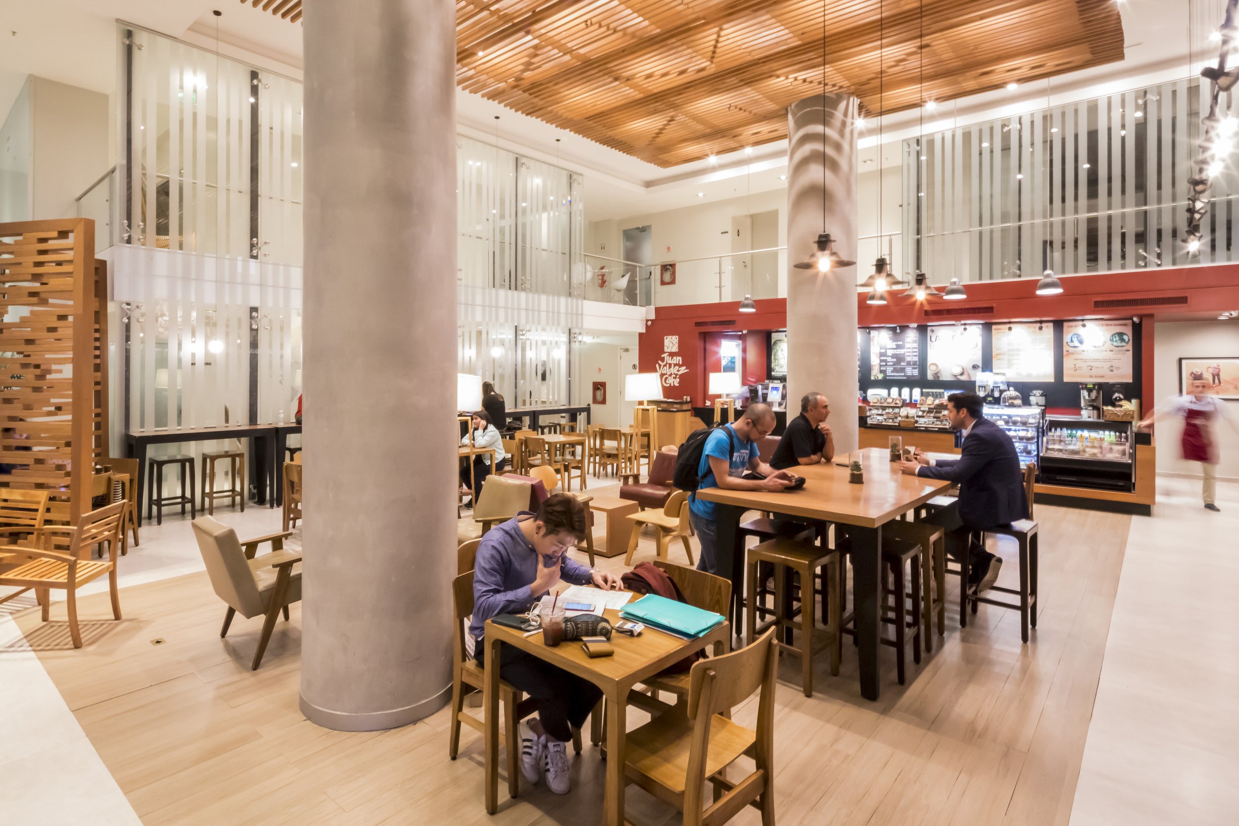 Interior de una cafetería moderna con varias personas sentadas en mesas y en la barra, paredes blancas, columna grande en el centro, y decoración en madera