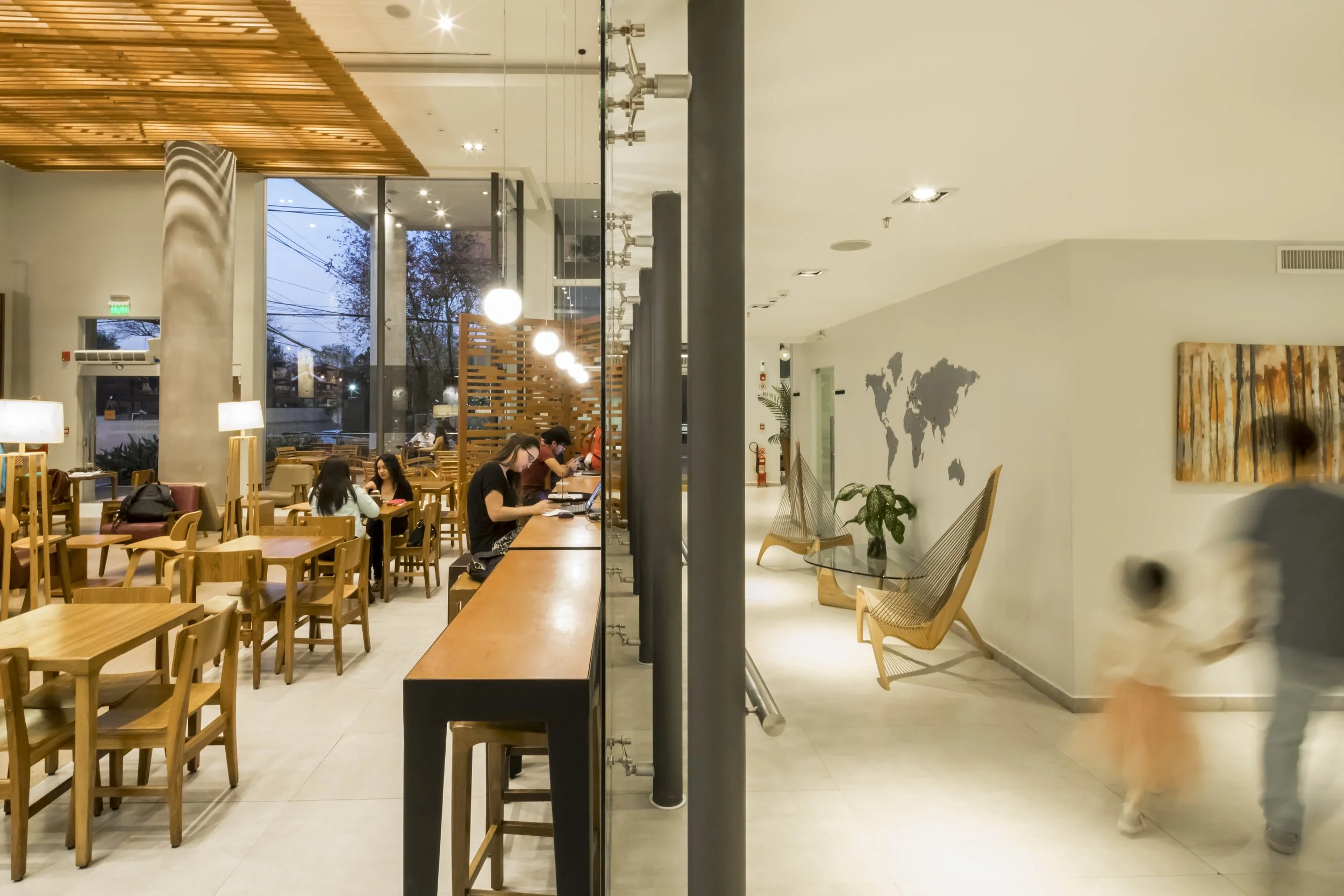 Interior de una cafetería con mesas de madera, sillas y una zona de computadoras donde algunas personas trabajan, paredes decoradas con un mapa del mundo y un cuadro abstracto, luz cálida, vista hacia la calle