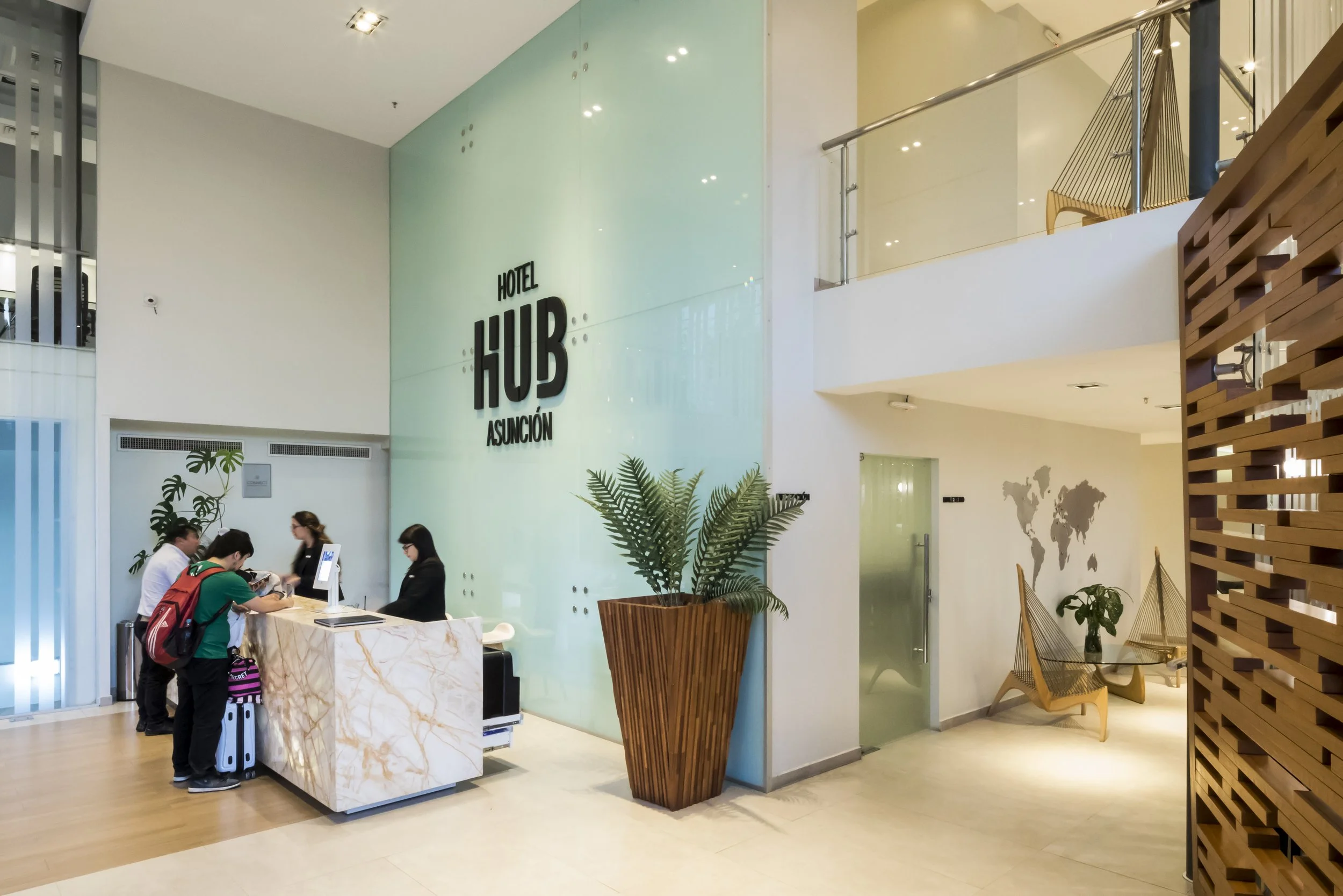Recepción del Hotel Hub Asunción con personas registrándose y ambiente moderno con plantas y decoraciones de madera.