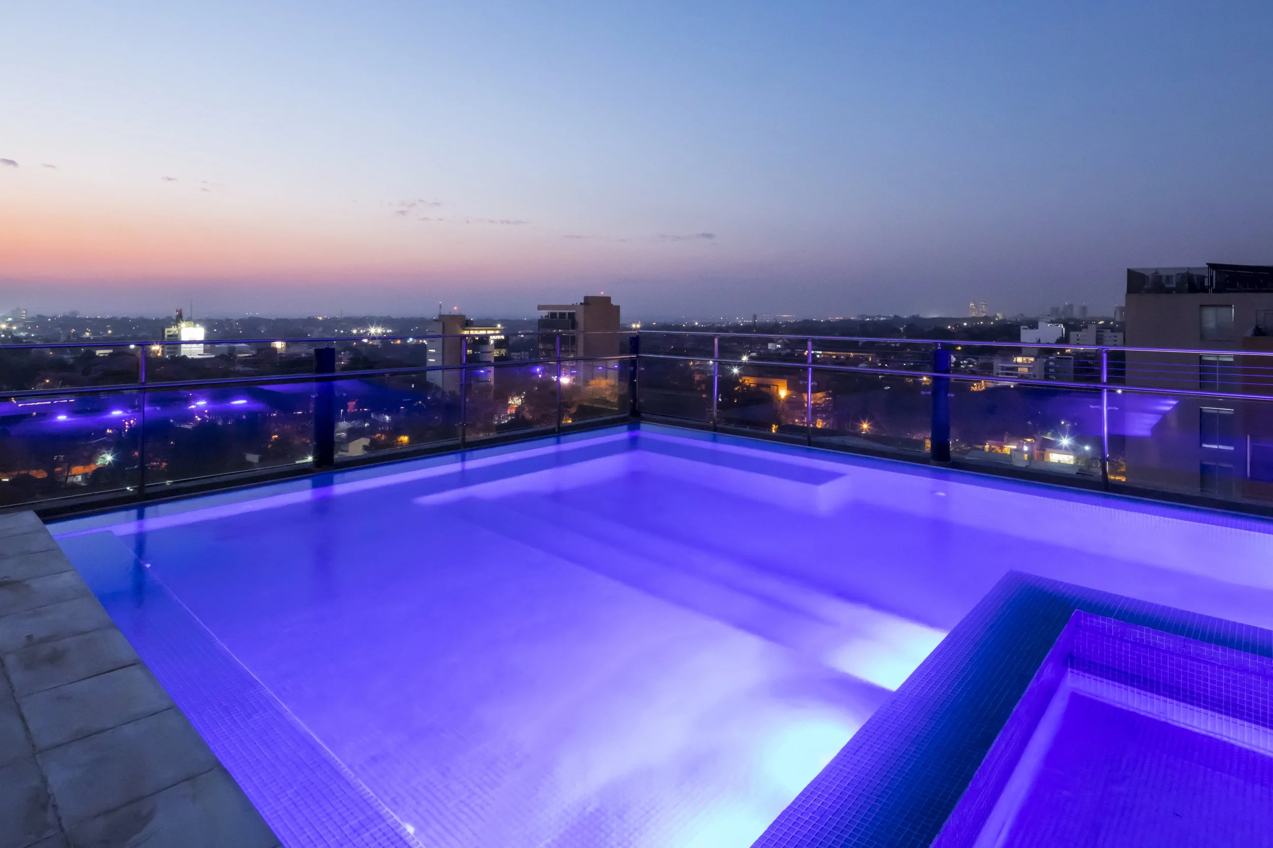 Piscina en la azotea con iluminación azul, vista nocturna de la ciudad y cielo al atardecer