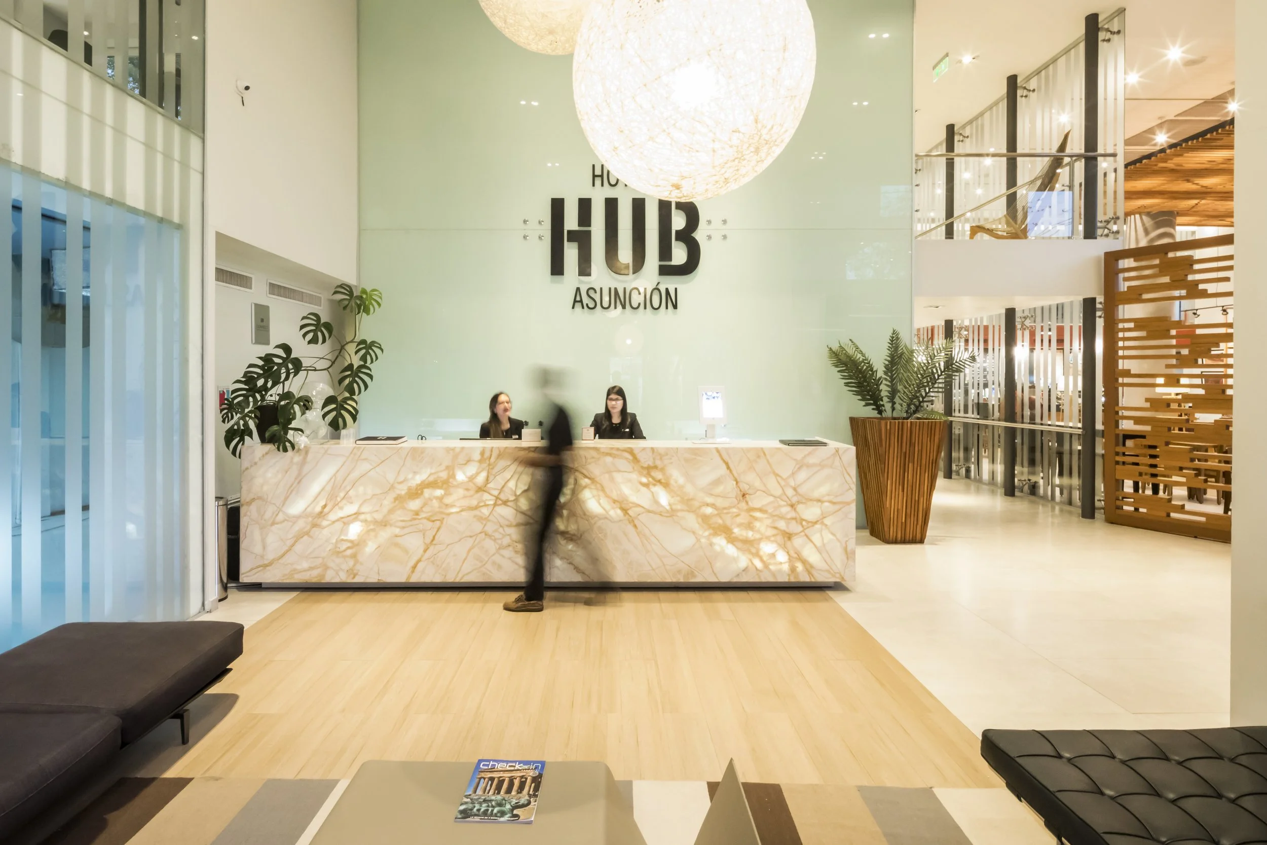 Recepción del Hotel HUB Asunción, con un mostrador de mármol, dos recepcionistas y una planta en una esquina, en un ambiente moderno y bien iluminado.