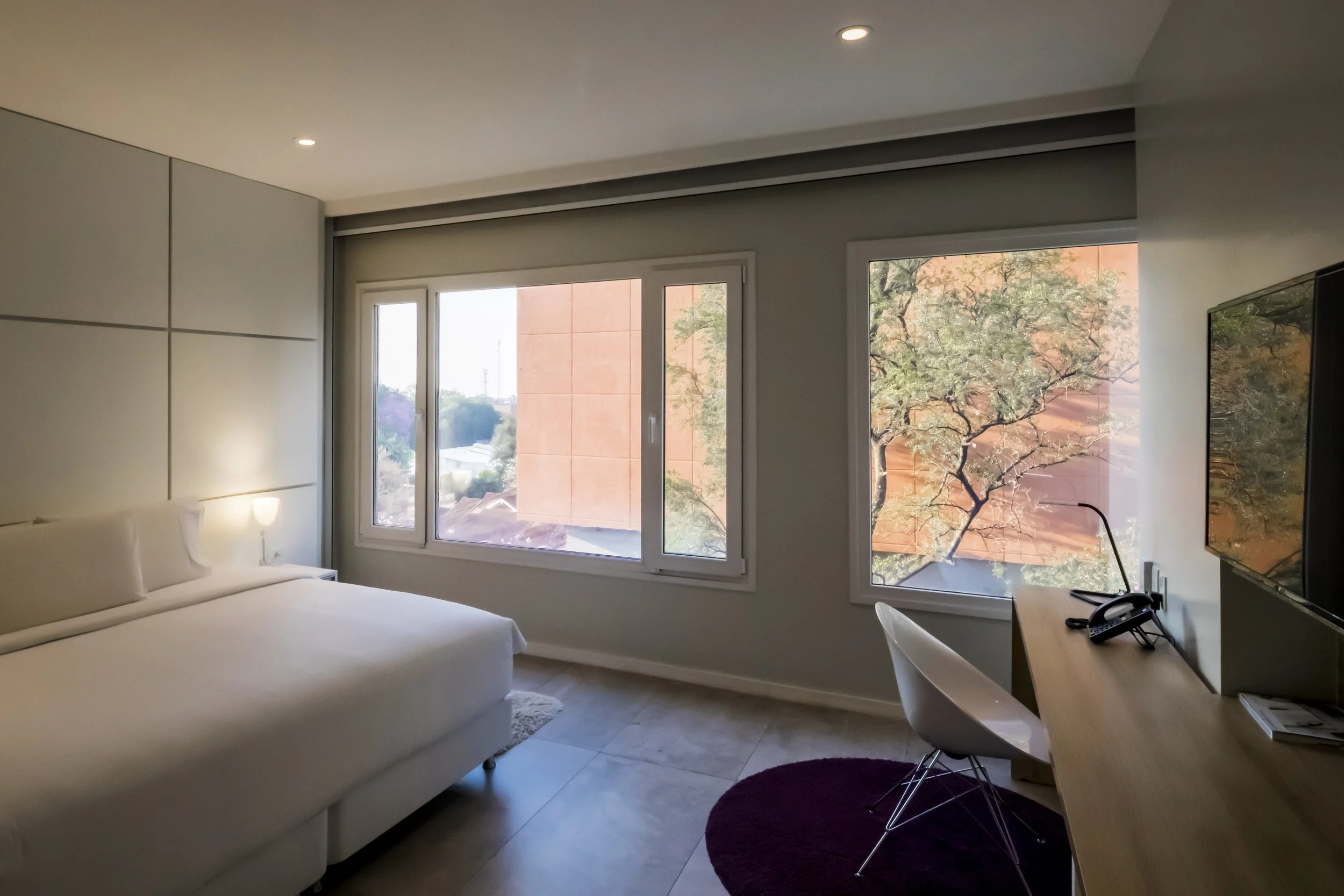 Habitación moderna con una cama blanca, ventanas grandes con vista a árboles y edificios, escritorio con teléfono y silla, con iluminación suave y piso de baldosas claras.