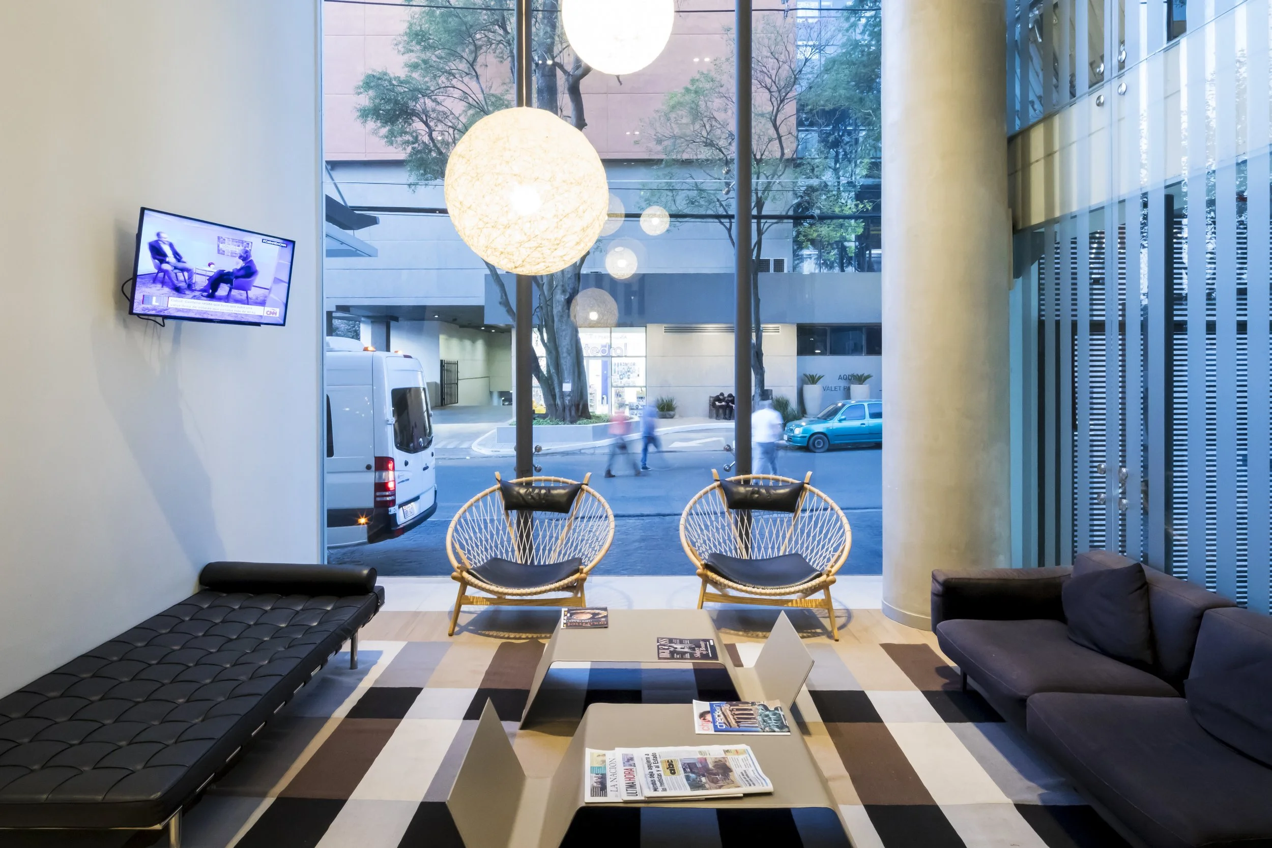 Sala de espera moderna con sillones de mimbre y malla, una televisión en la pared, mesas con revistas y un ventanal grande con vista a la calle con autos y personas paseando.