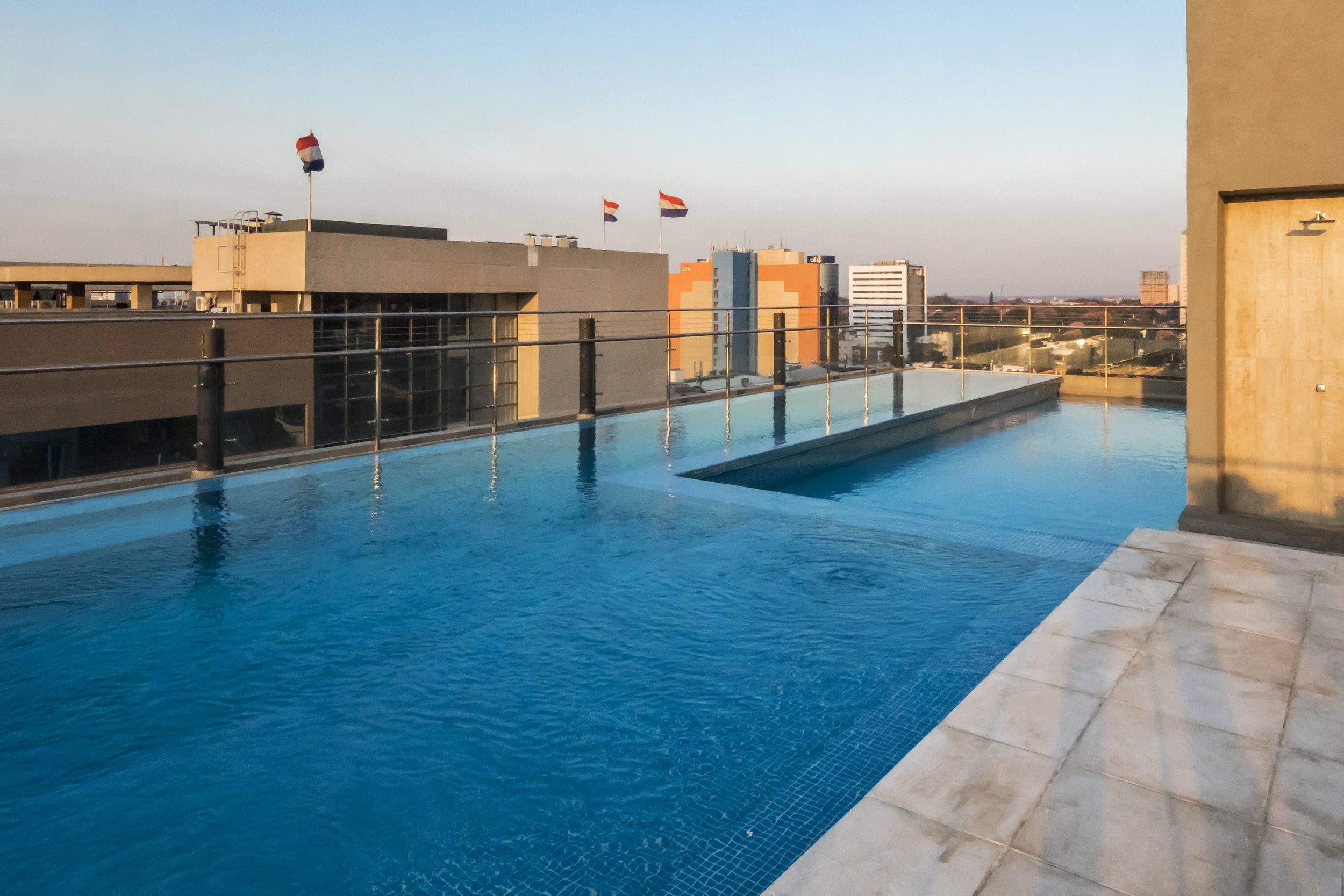 Piscina de azulejos azules en la azotea de un edificio en la ciudad, con banderas nacionales ondeando y vista a otros edificios y el horizonte.