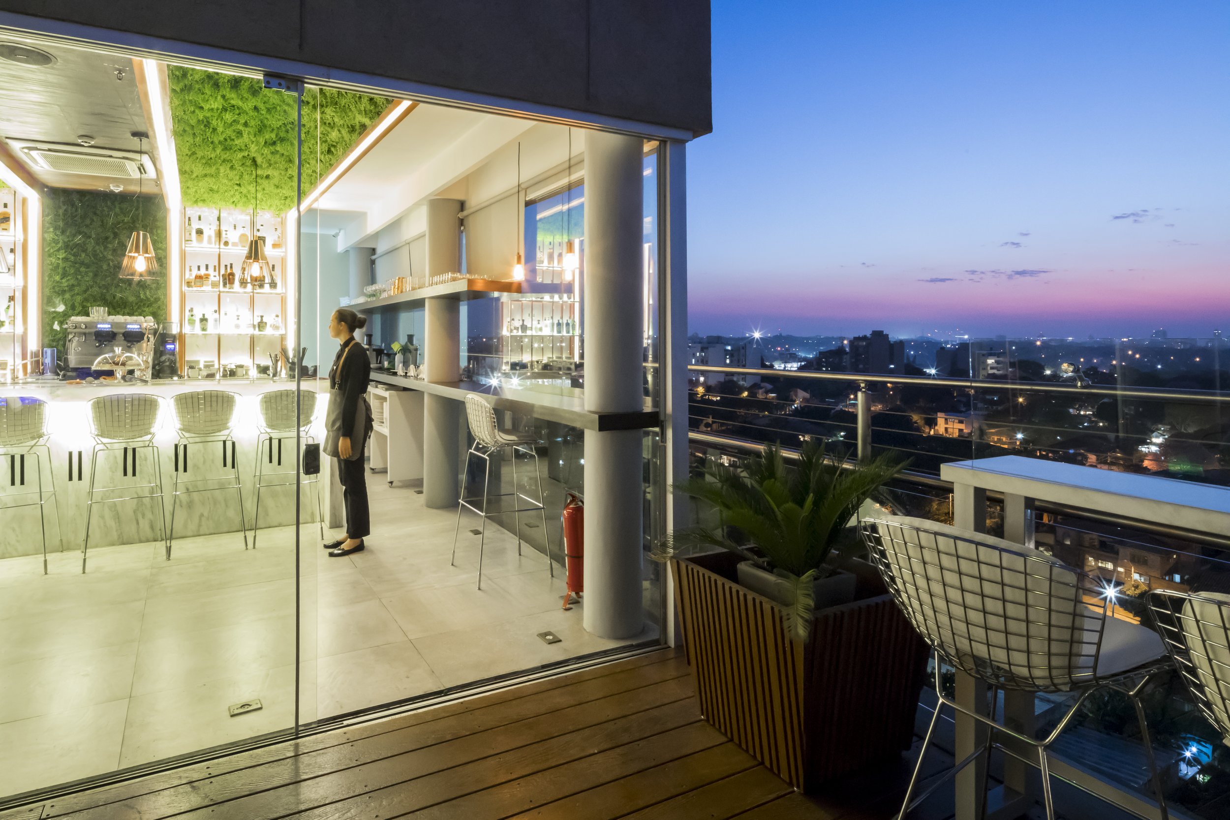 Vista de un bar en un edificio alto durante el atardecer, con una camarera de pie frente a la barra y vista panorámica de la ciudad y el cielo al atardecer.
