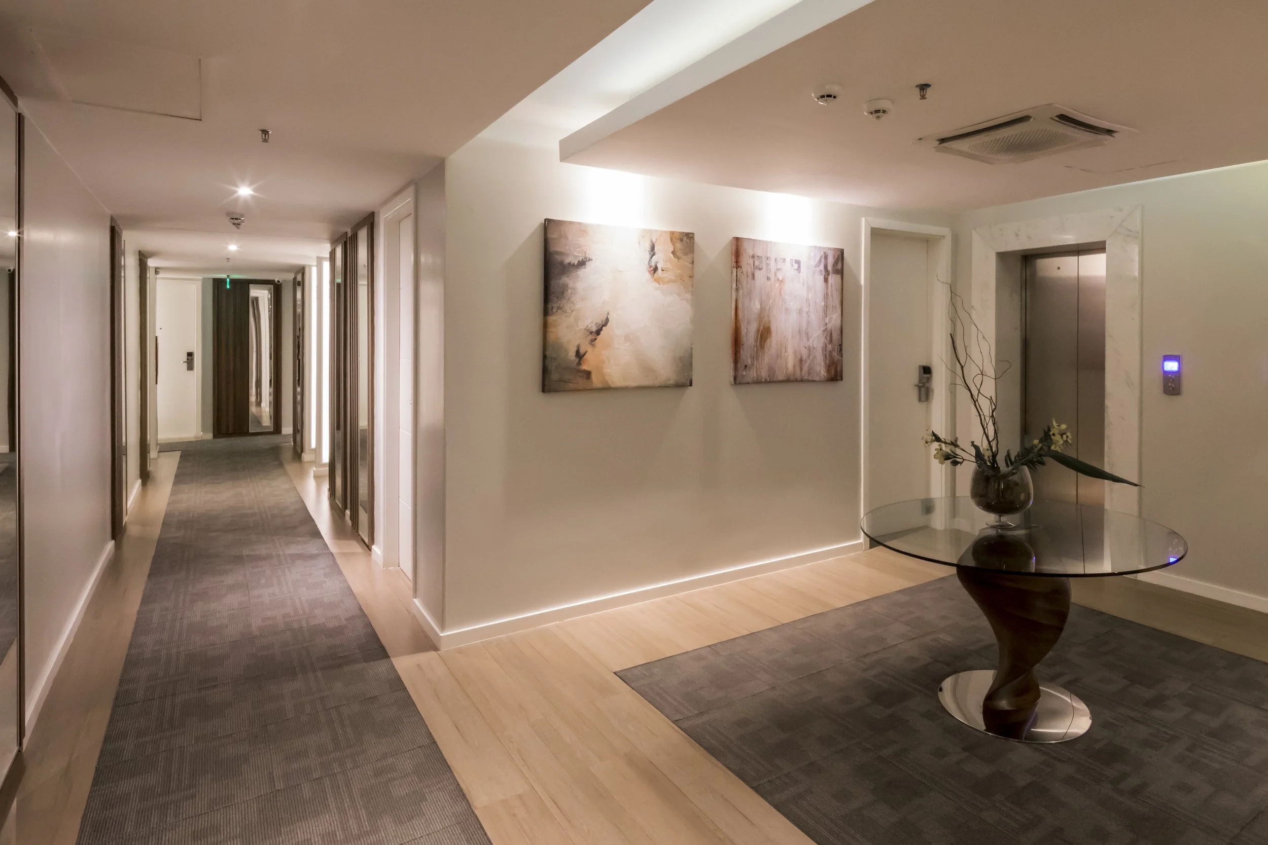 Vestíbulo de hotel con pasillo, dos cuadros abstractos en la pared, mesa redonda con florero y elevador