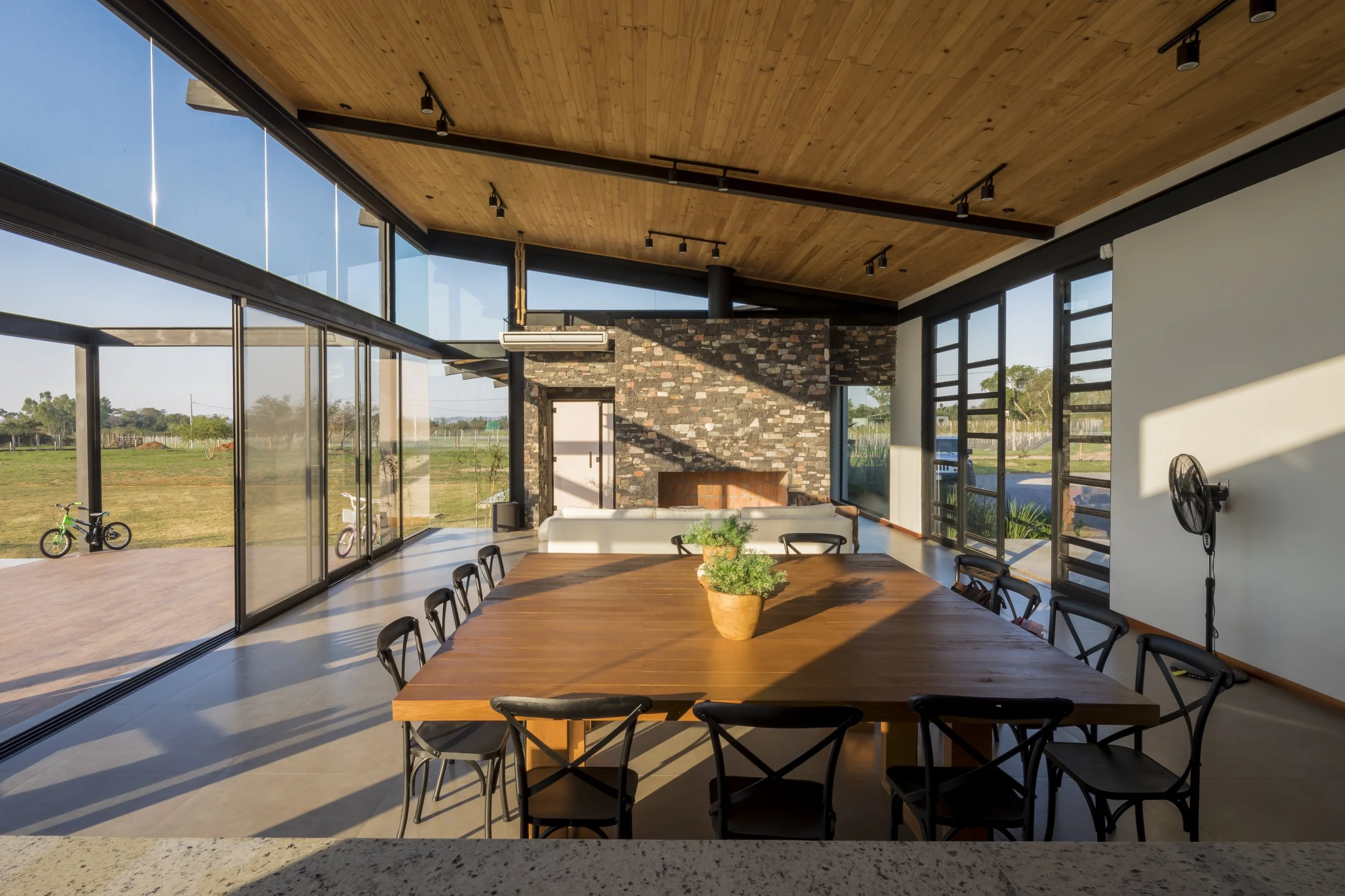 Interior moderno con mesa de madera y sillas negras, paredes de vidrio que muestran un campo exterior, chimenea de piedra y detalles en madera en el techo.