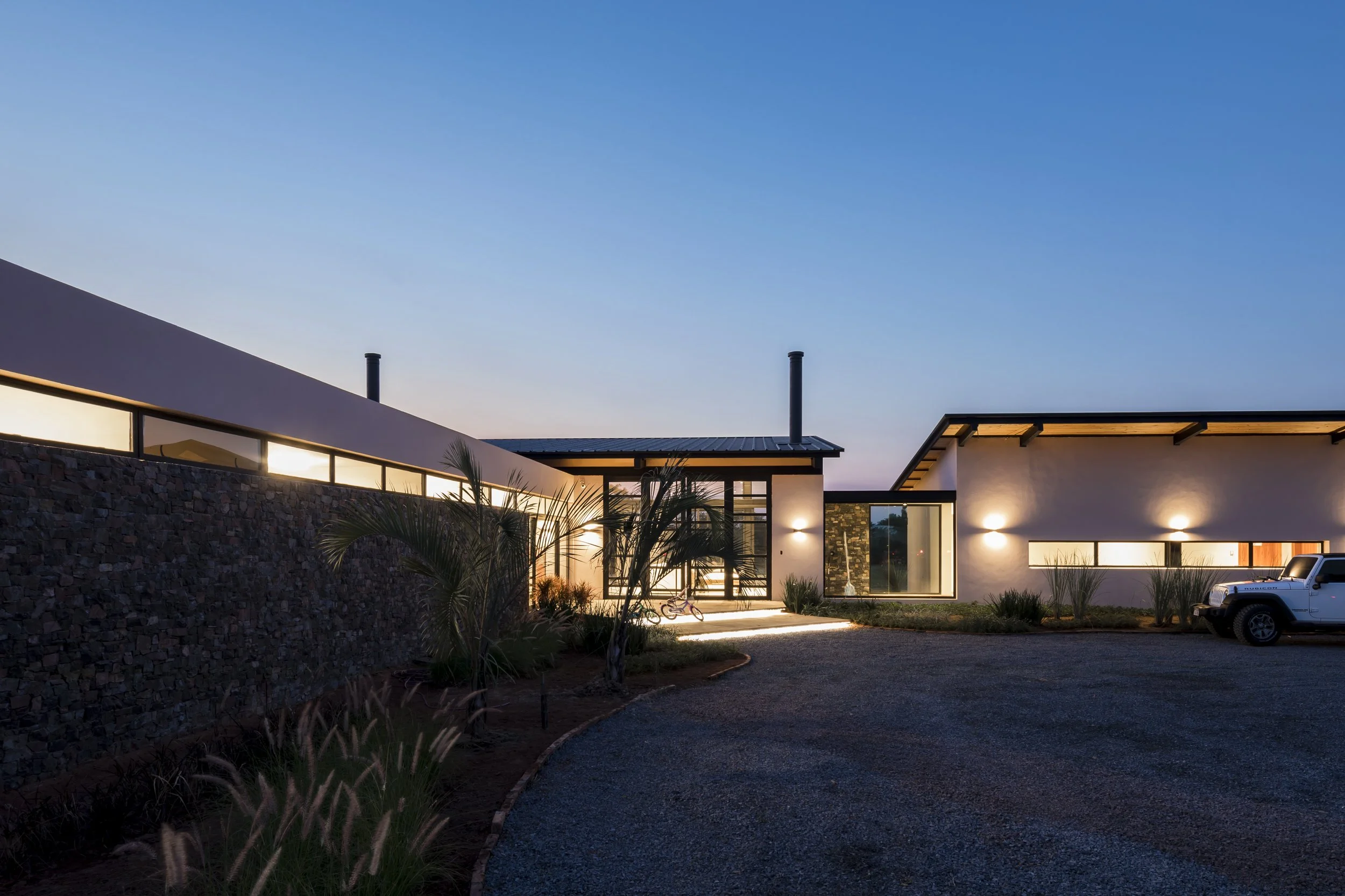 Casa moderna con muro de piedra, luces exteriores, árboles y coche, durante el atardecer.