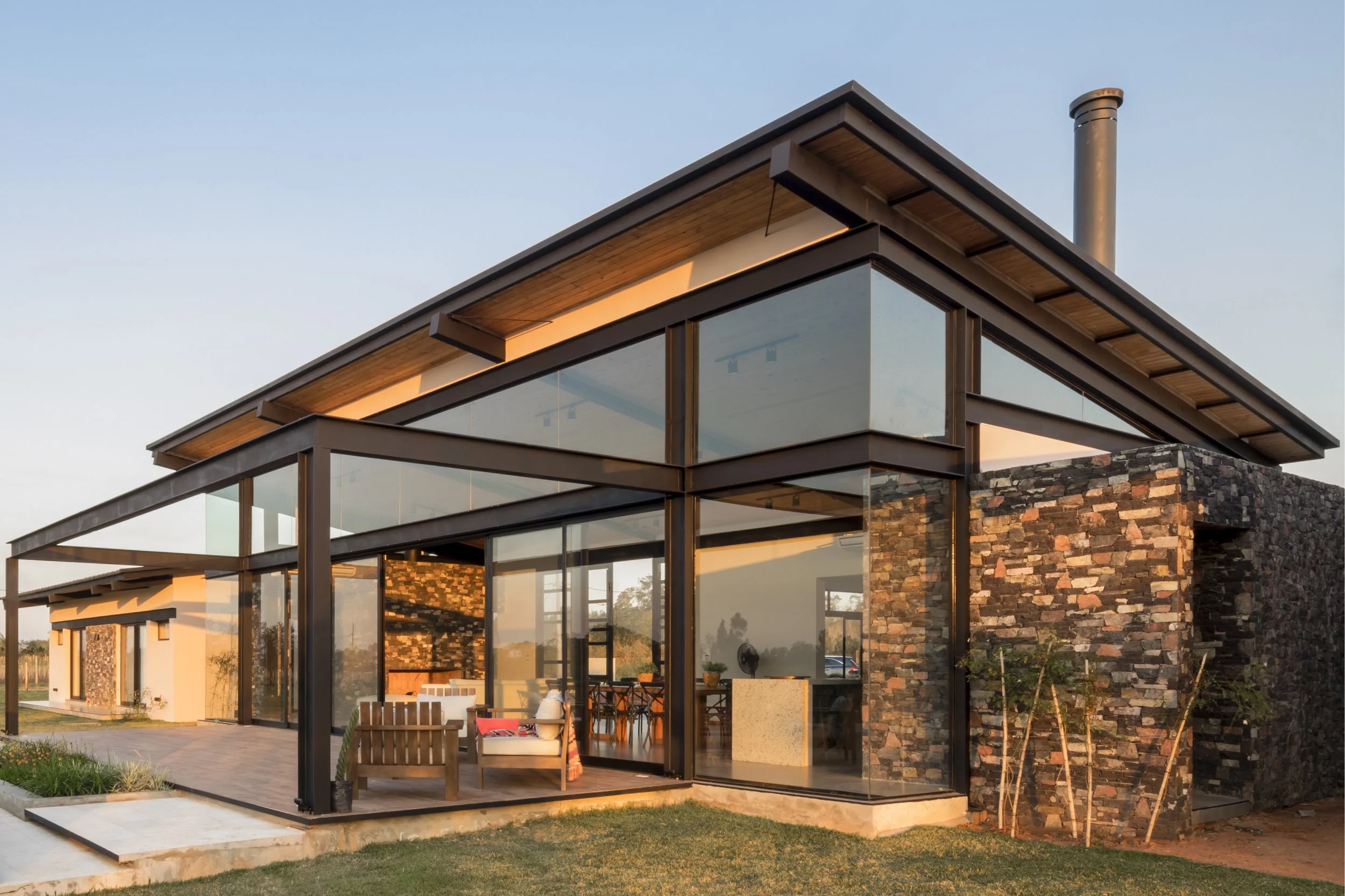 Casa moderna con amplio uso de cristalería, estructura de metal, paredes de piedra y área de terraza con sillones y mesa, al atardecer.