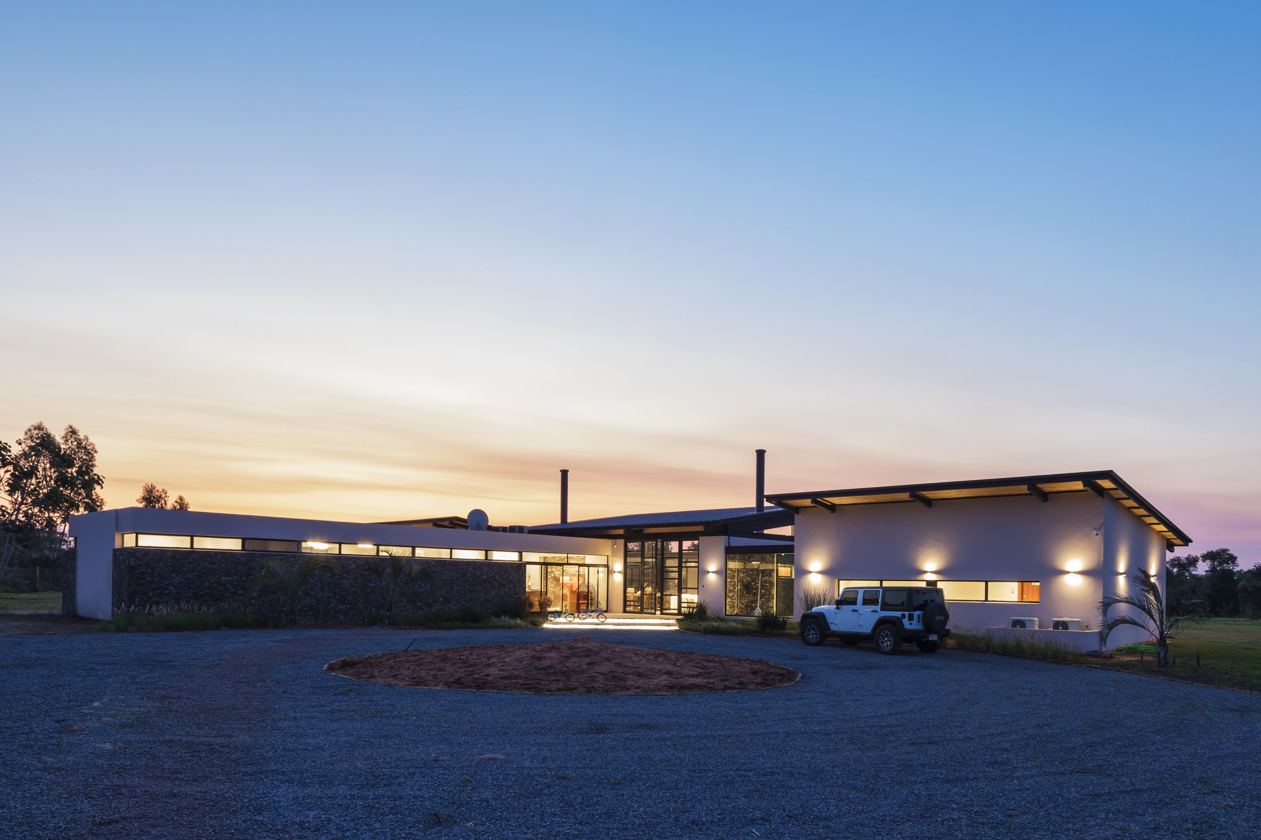 Casa moderna con luces en un atardecer, rodeada de un área de grava y algunos árboles en el fondo.