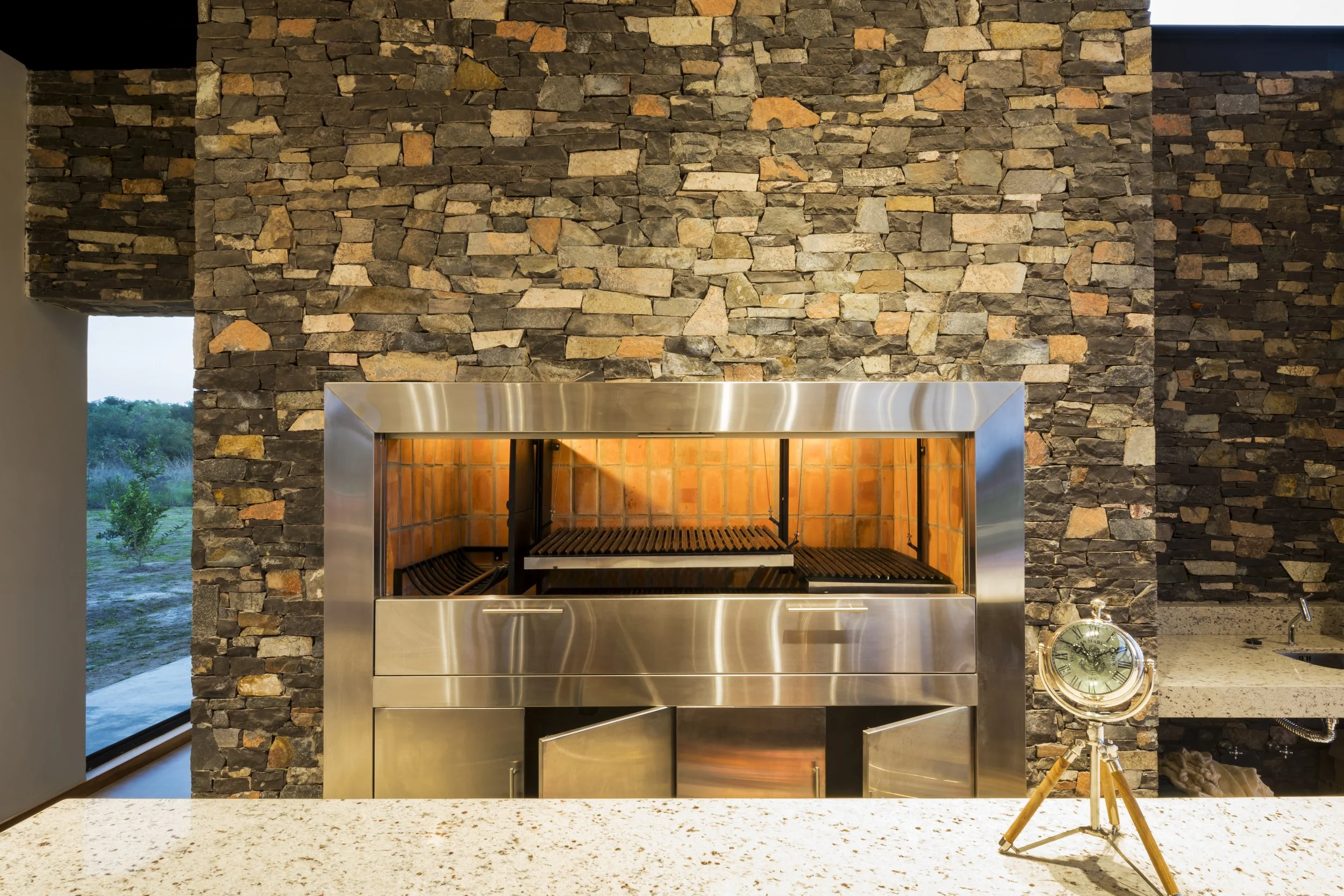Horno de barbacoa de acero inoxidable en una pared de piedra rústica, con un reloj de madera y latón en una encimera de piedra clara.