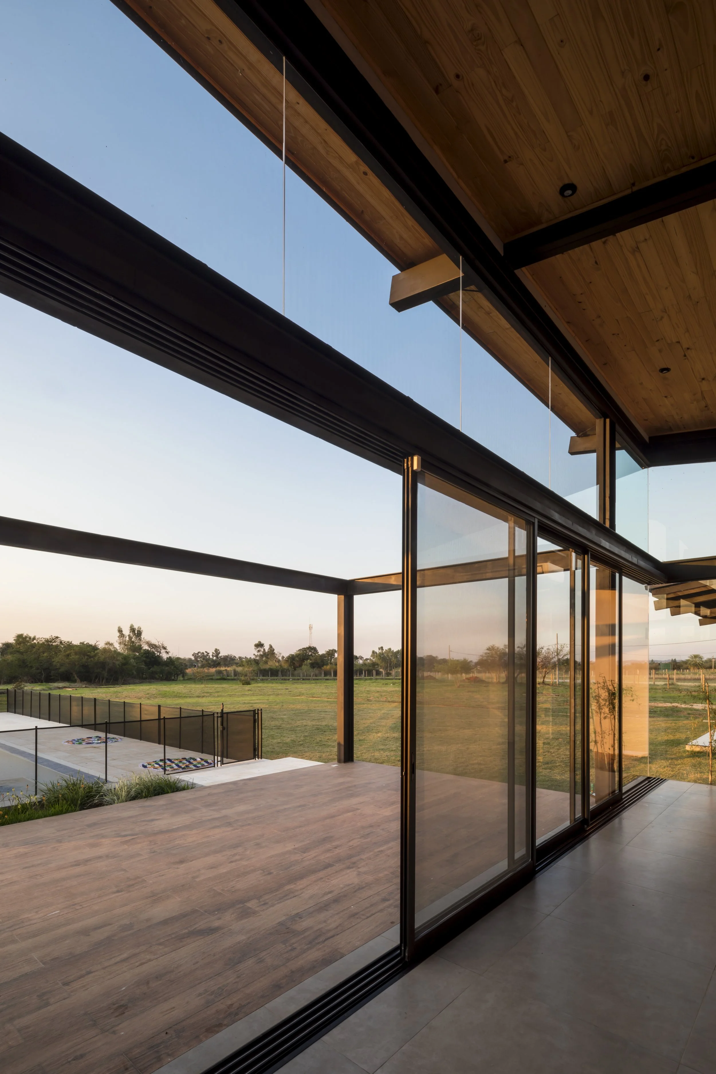 Vista desde el interior de una casa de diseño moderno con ventanas de vidrio que permiten mirar hacia el exterior, donde hay un patio y un campo con pasto, árboles y cielo despejado al atardecer.