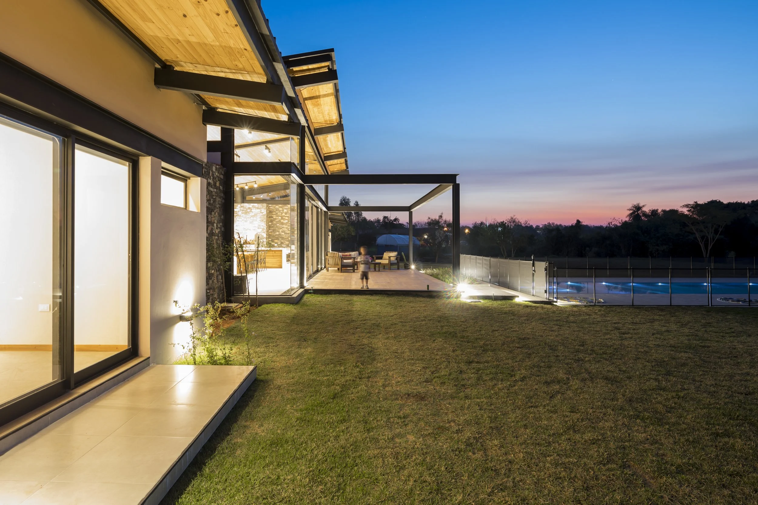 Casa moderna con ventanales de vidrio, patio con césped, área de descanso al aire libre, piscina y árboles al atardecer.