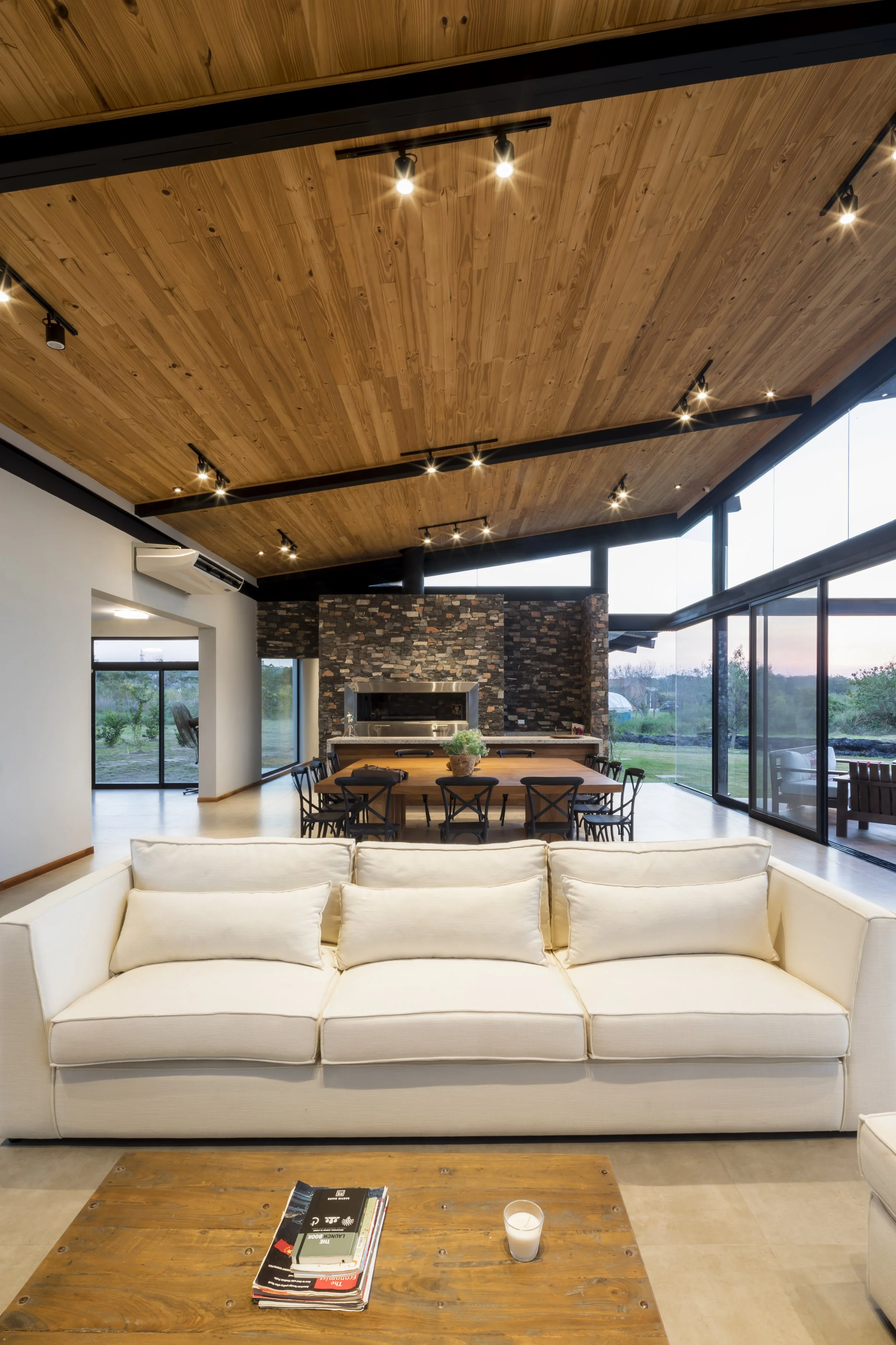 Sala de estar moderna con sillón blanco, mesa de centro de madera, chimenea de piedra, mesa de comedor con sillas negras y gran ventanal con vista al exterior.