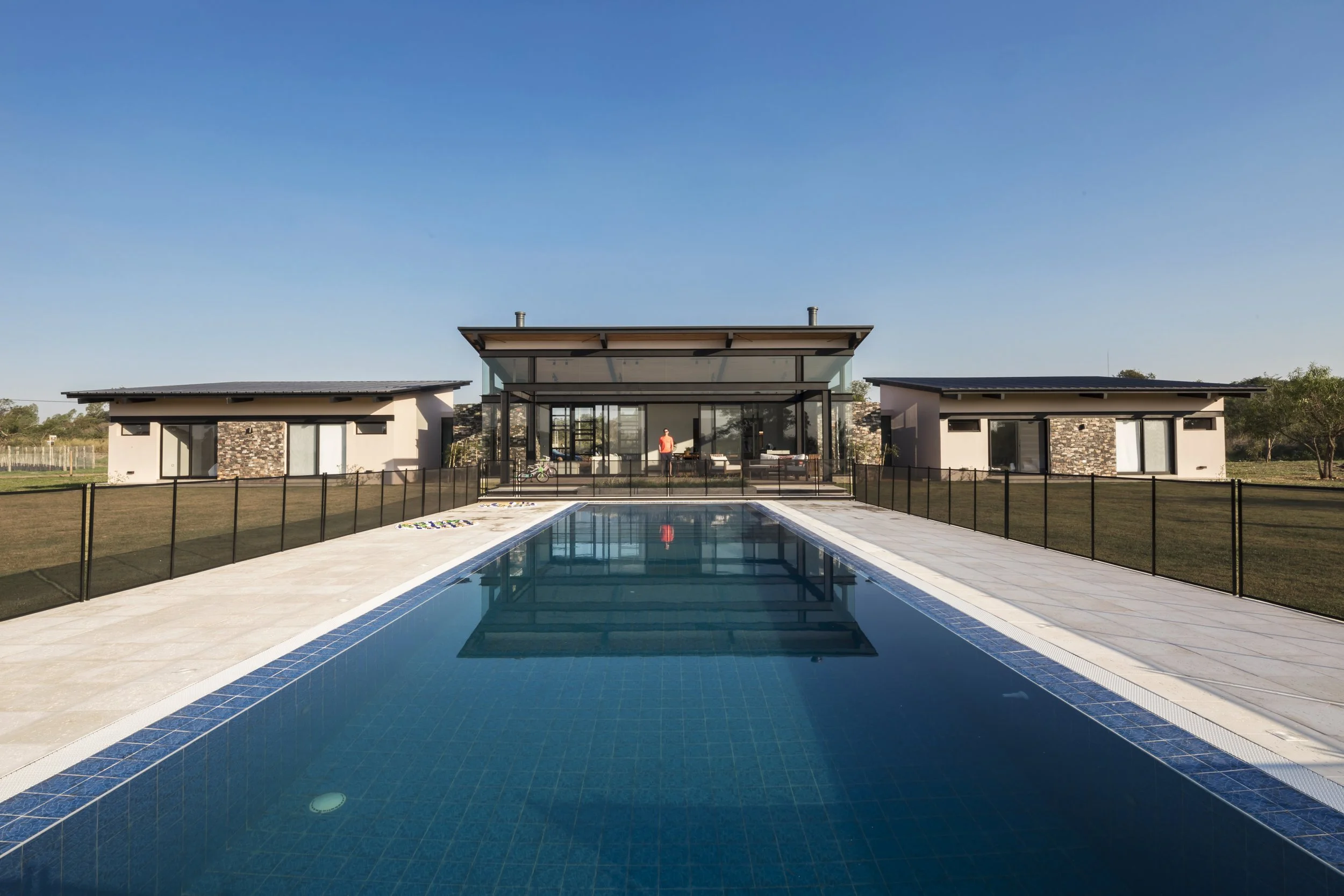 Casa moderna con piscina y amplias ventanas, rodeada de jardín y césped bajo cielo despejado.